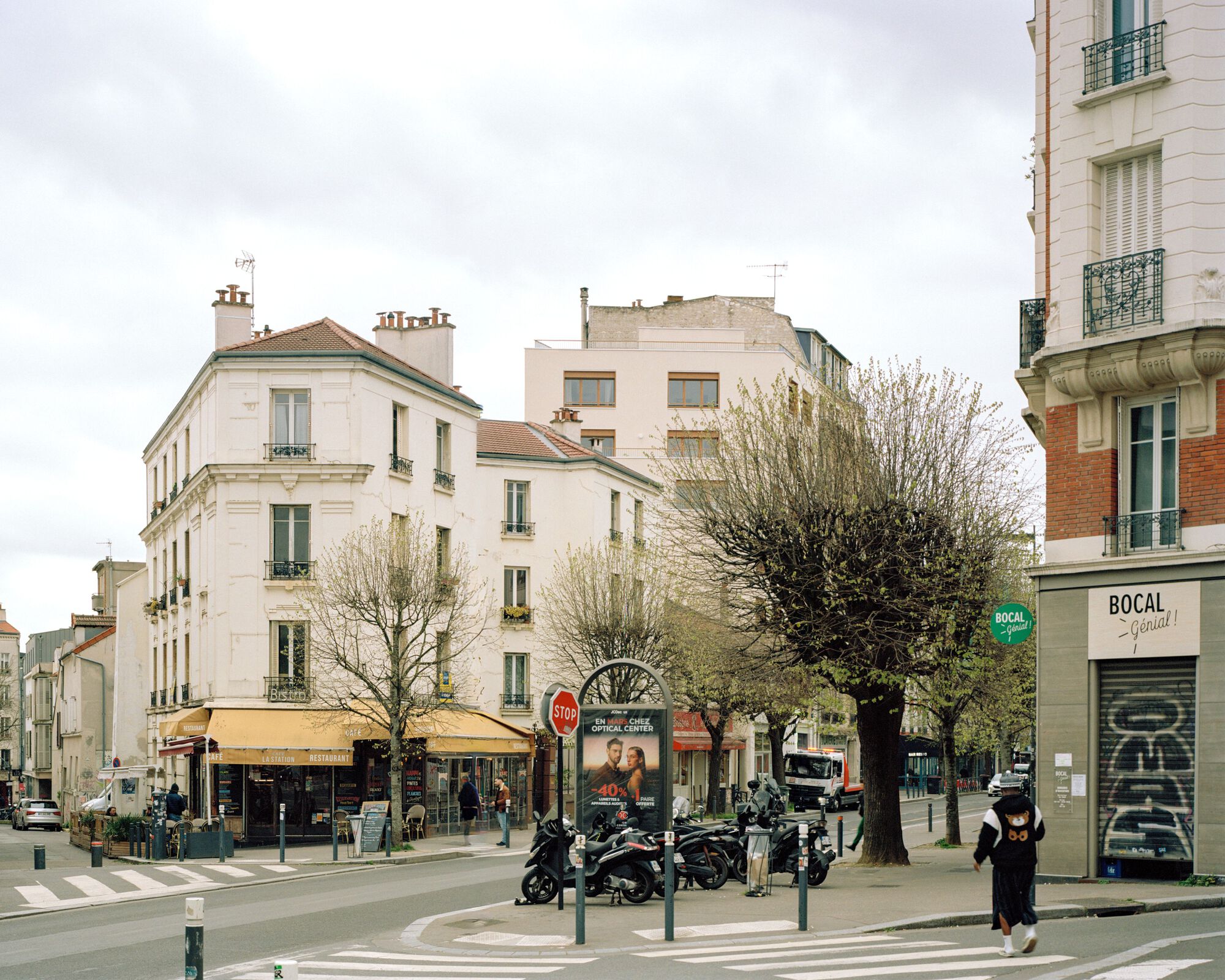 Street corner of cream stone apartment buildings with an awning, pollarded trees and parked scooters, a pedestrian crossing in overcast spring light