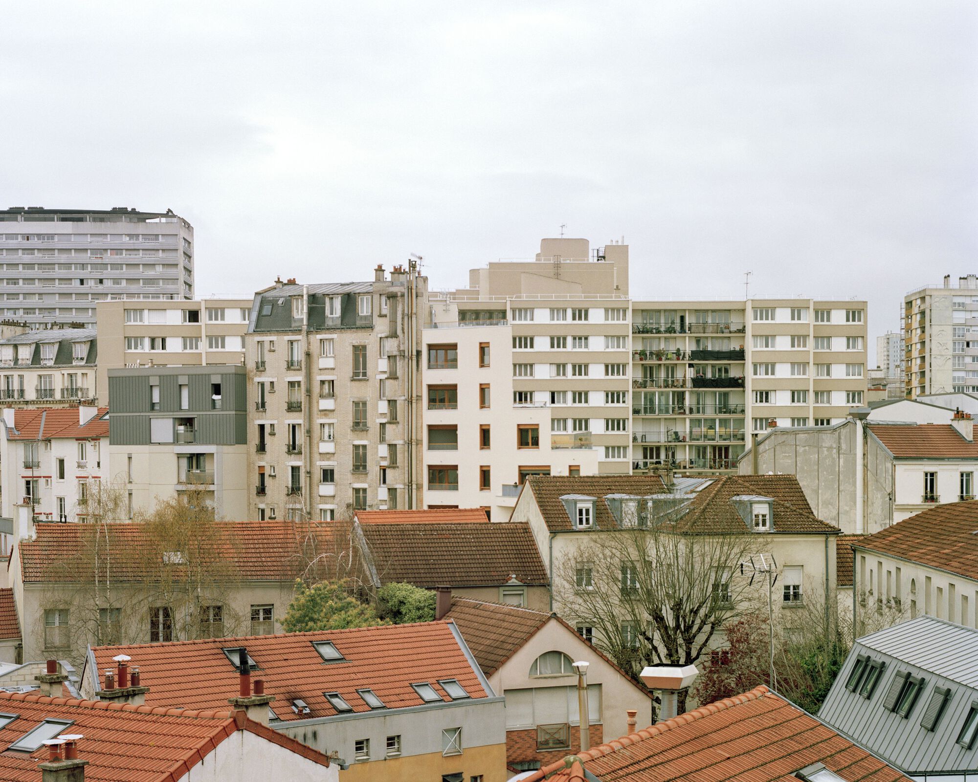 Rooftop view over terracotta tile roofs toward mid-rise apartment blocks and bare trees under a flat grey sky