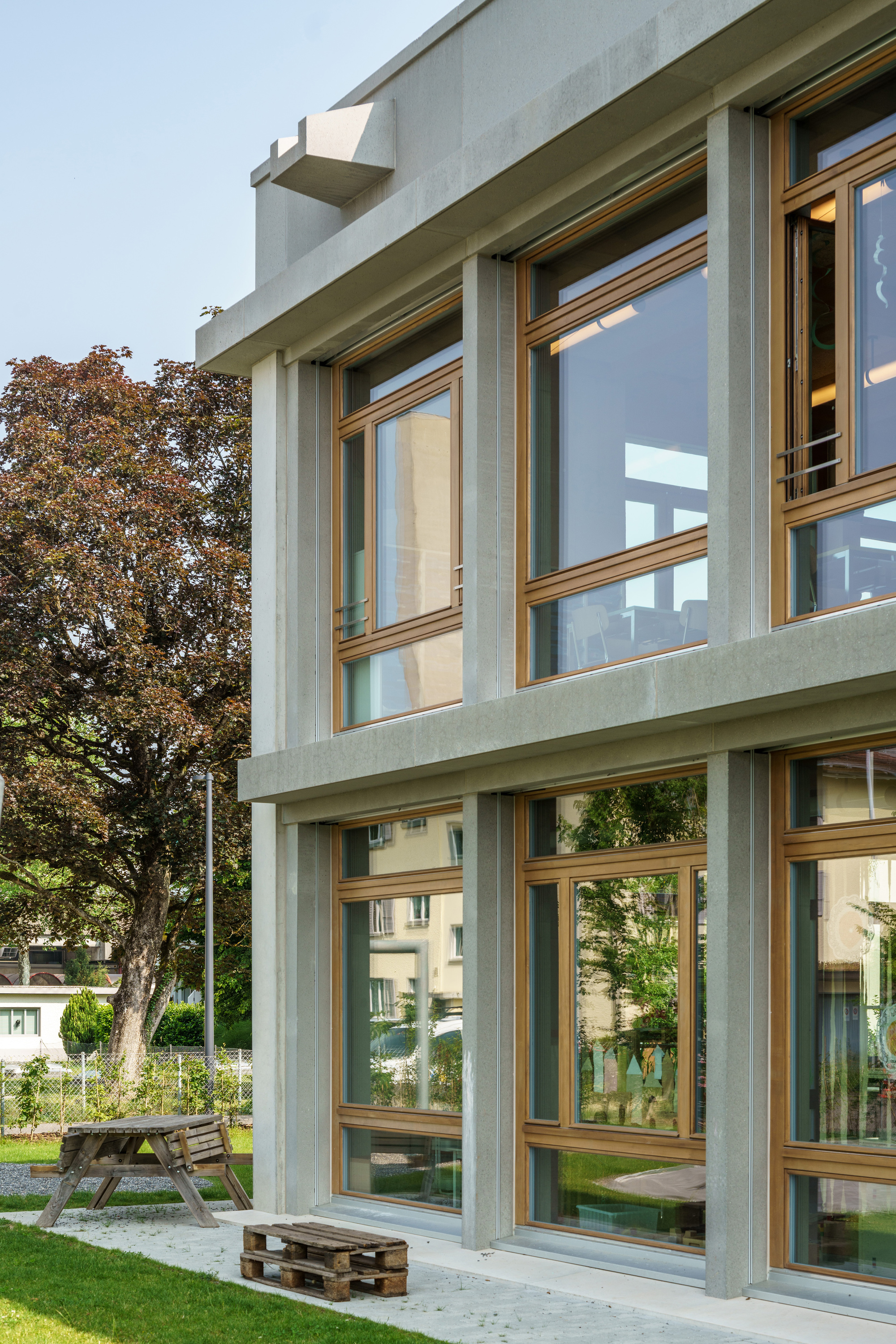 Close view of precast concrete columns and timber-framed glazing, with a picnic table and pallet seat on the lawn