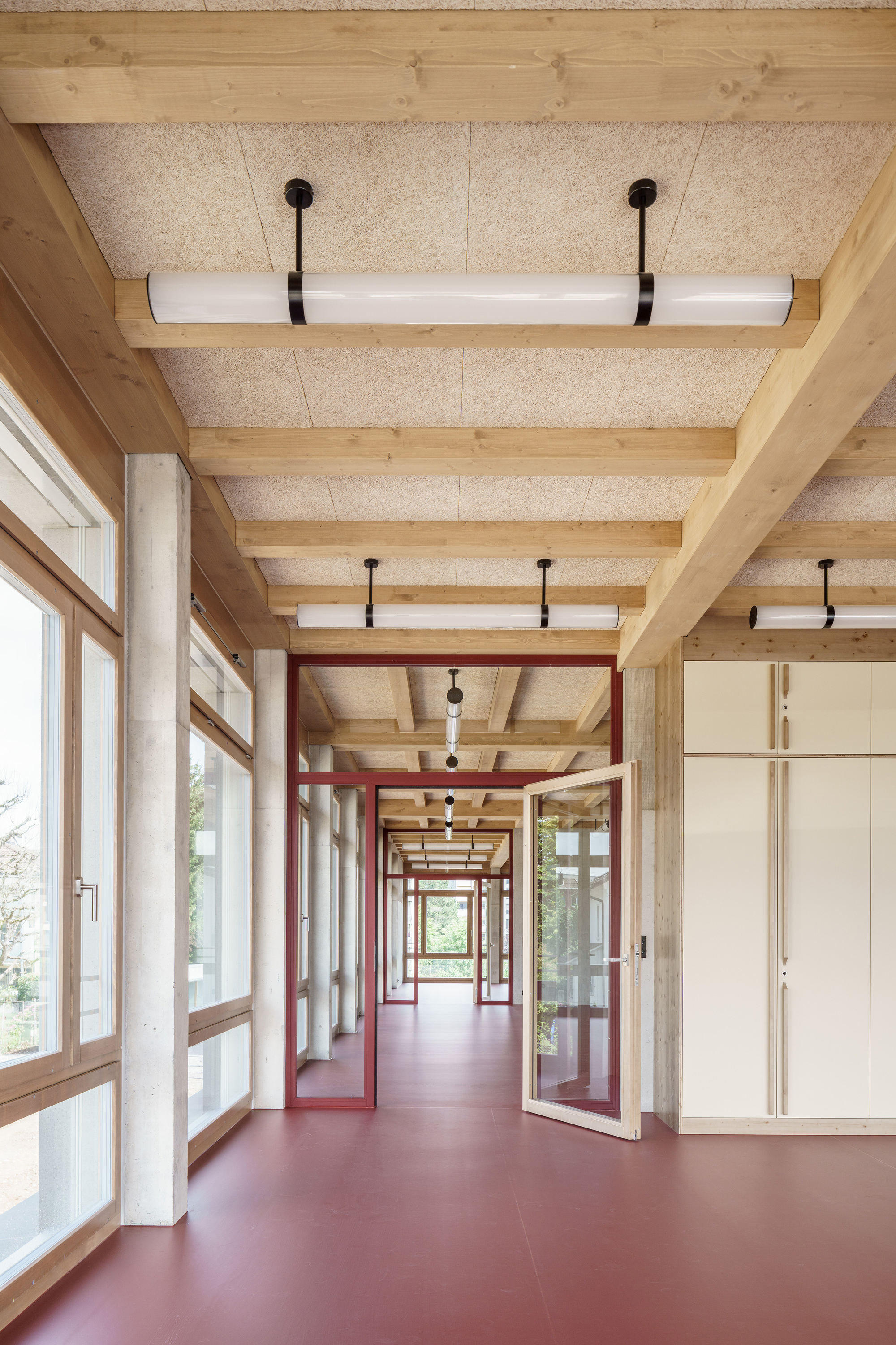 Corridor with exposed timber beams, tube pendant lights, red-framed glass doors and a red floor, daylight entering from the left