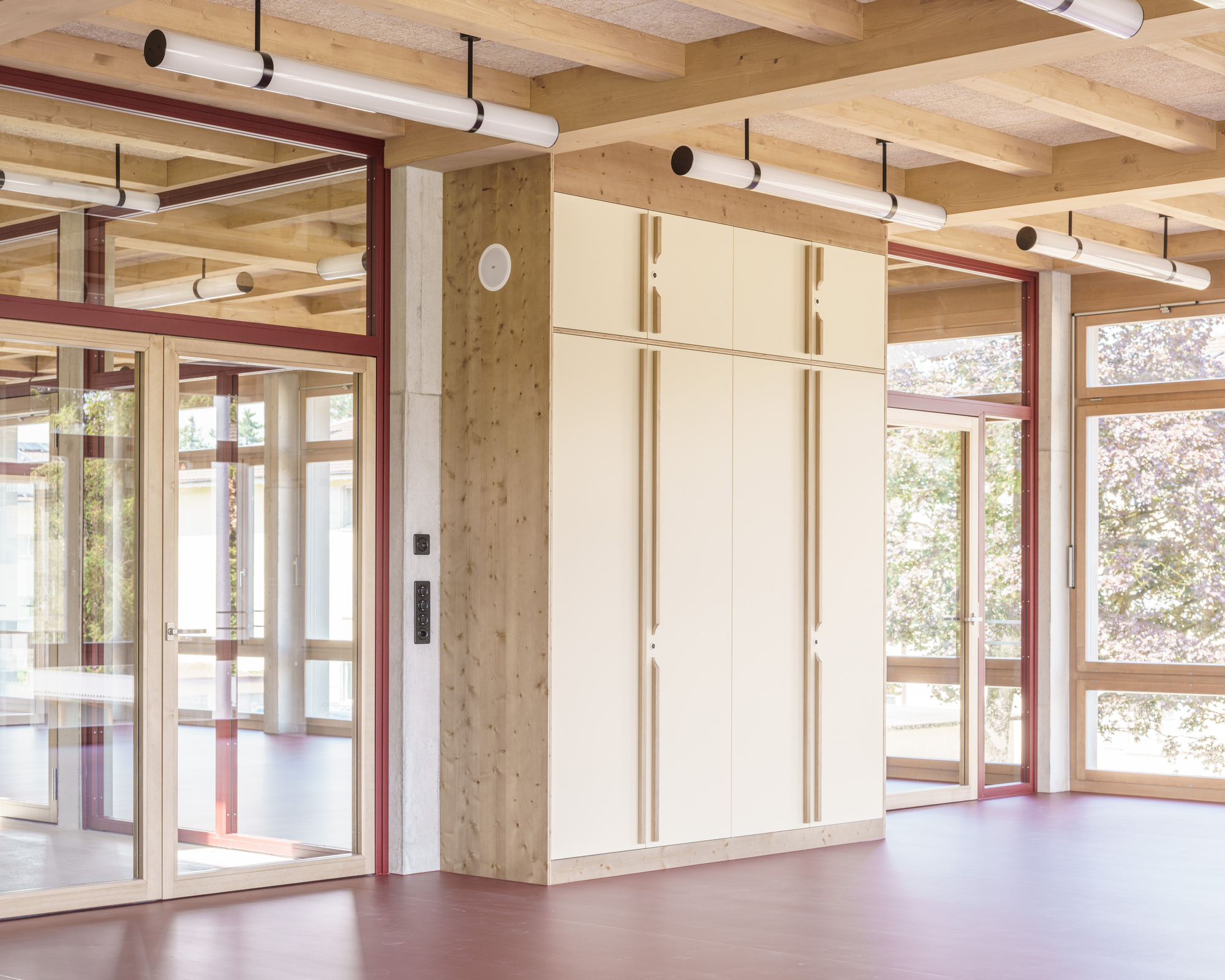 Open room with cream cabinet block, timber column, red-framed glazed doors and pendant tube lights, with a leafy tree outside