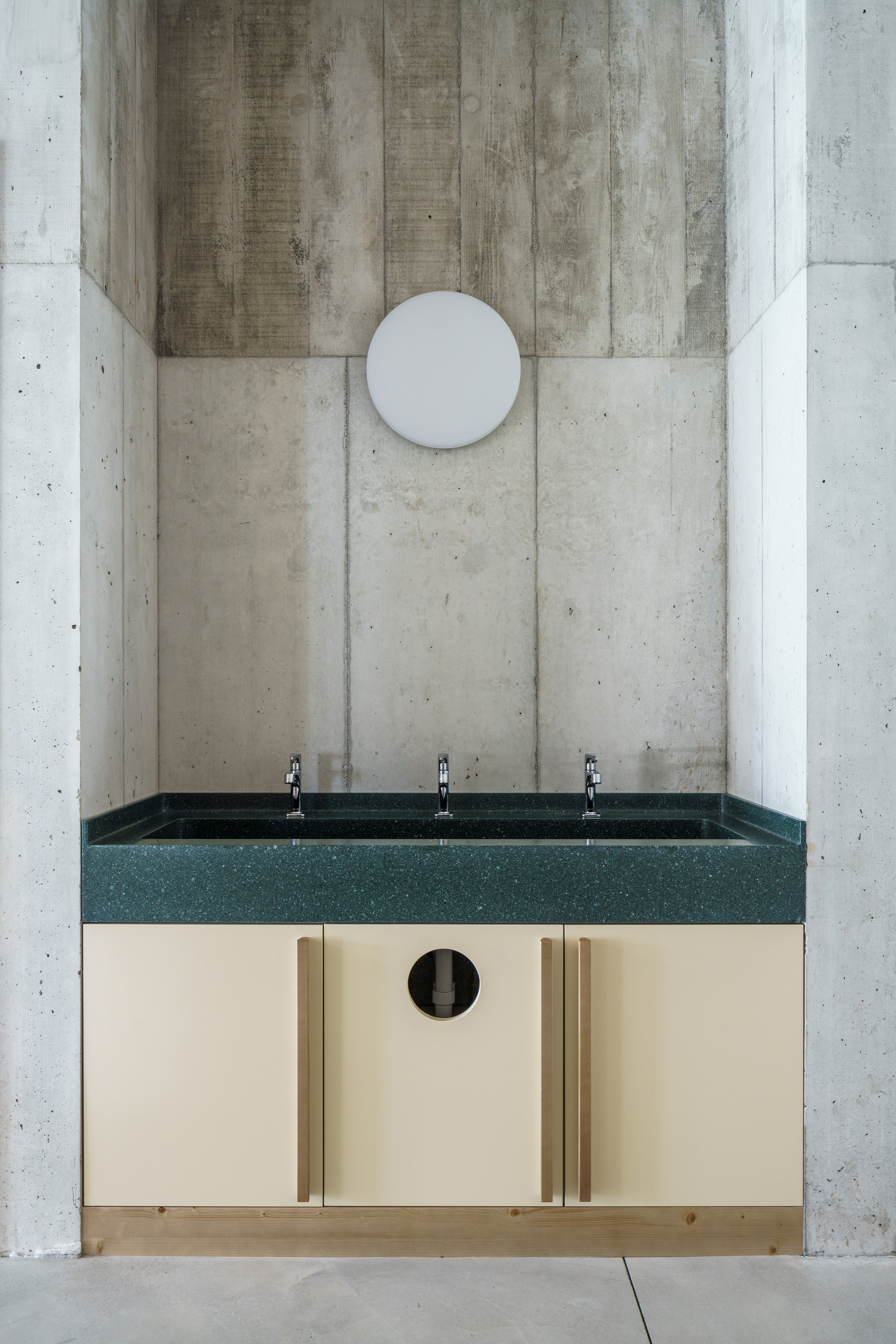 Three-tap washbasin with green speckled top and cream cabinet doors set in a board-marked concrete alcove under a round wall light