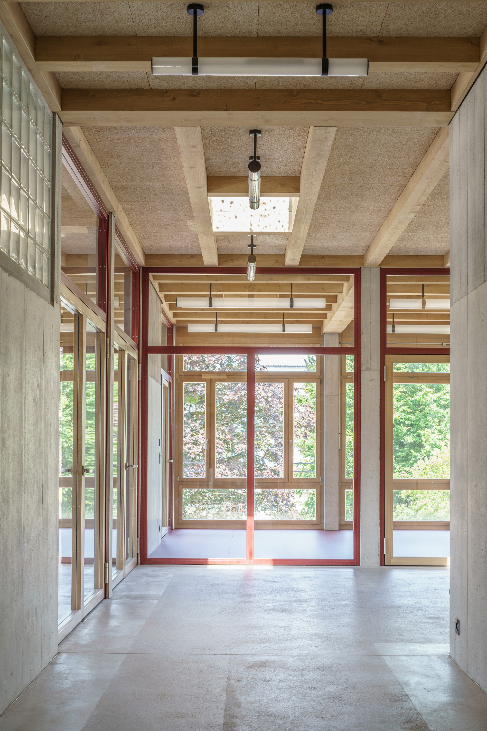 Corridor with exposed timber beams, red steel-framed glazing and a glass block wall, opening to green trees beyond