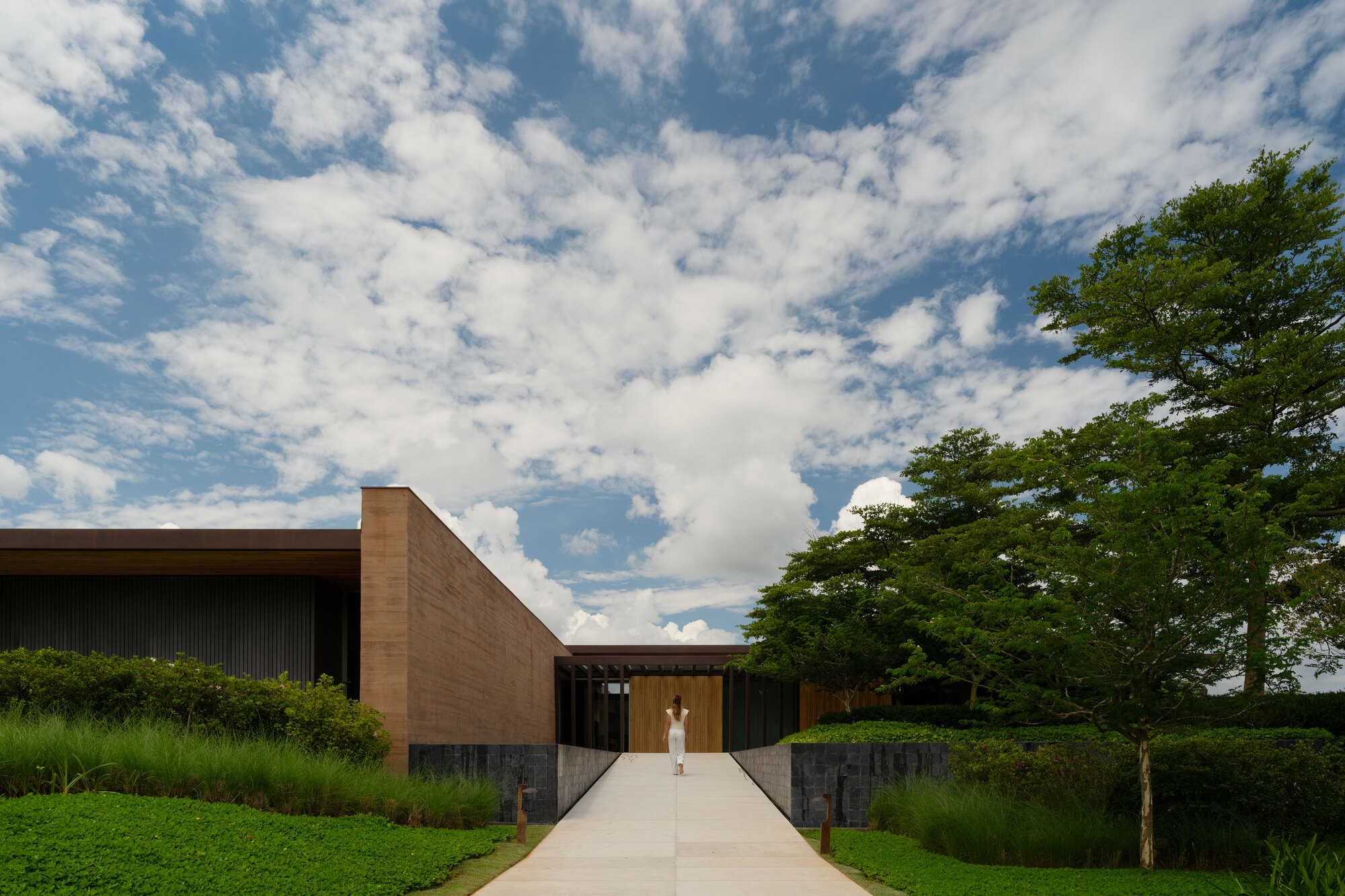 Concrete path leading to a low pavilion with a tall rammed earth wall, a woman in white walking under scattered clouds