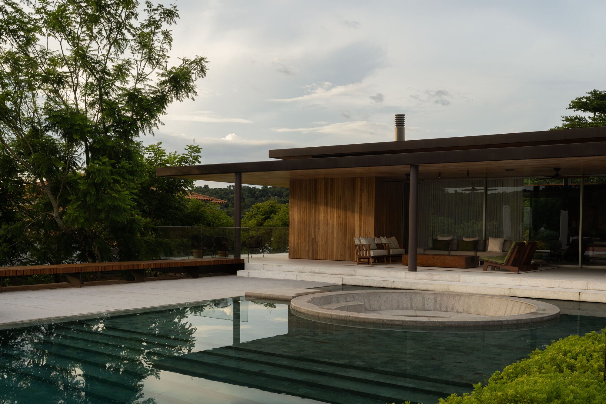 Pool with a circular sunken seating area in front of a timber-clad pavilion, seen in soft evening light