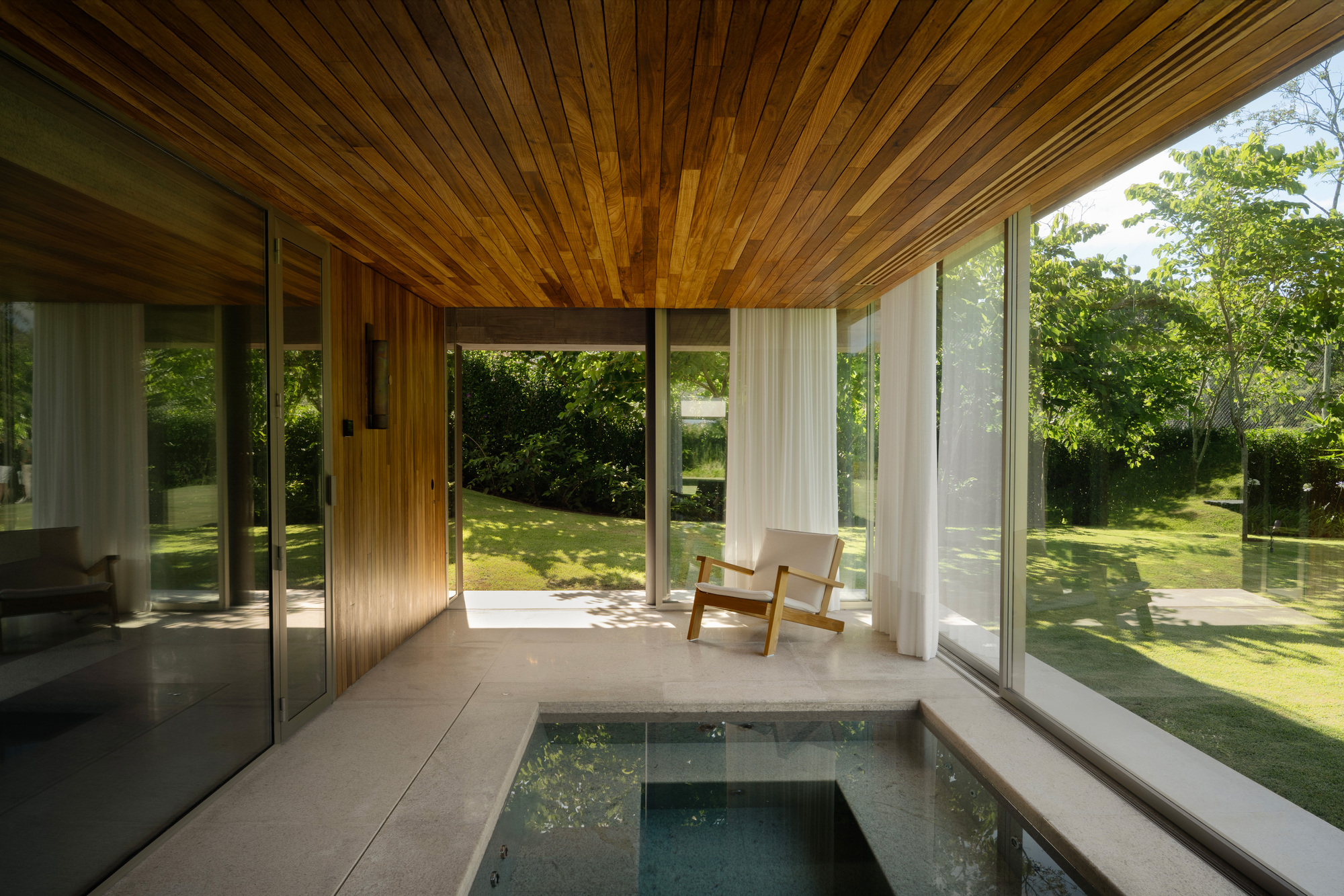 Covered terrace with a teak slat ceiling, a small plunge pool and a lounge chair beside sliding glass walls and sunlit lawn