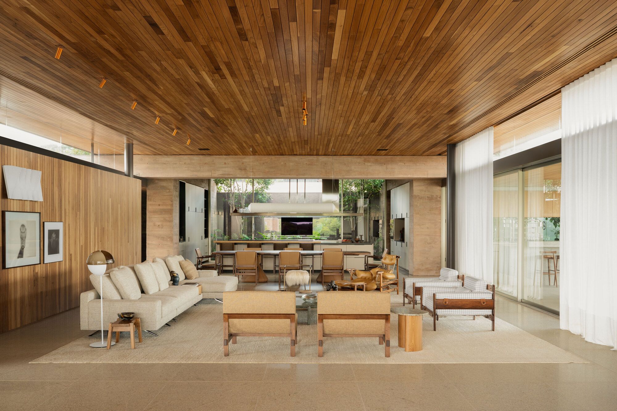 Open living room with a timber slat ceiling, low sofas and armchairs on a wool rug, and white curtains along the glazing