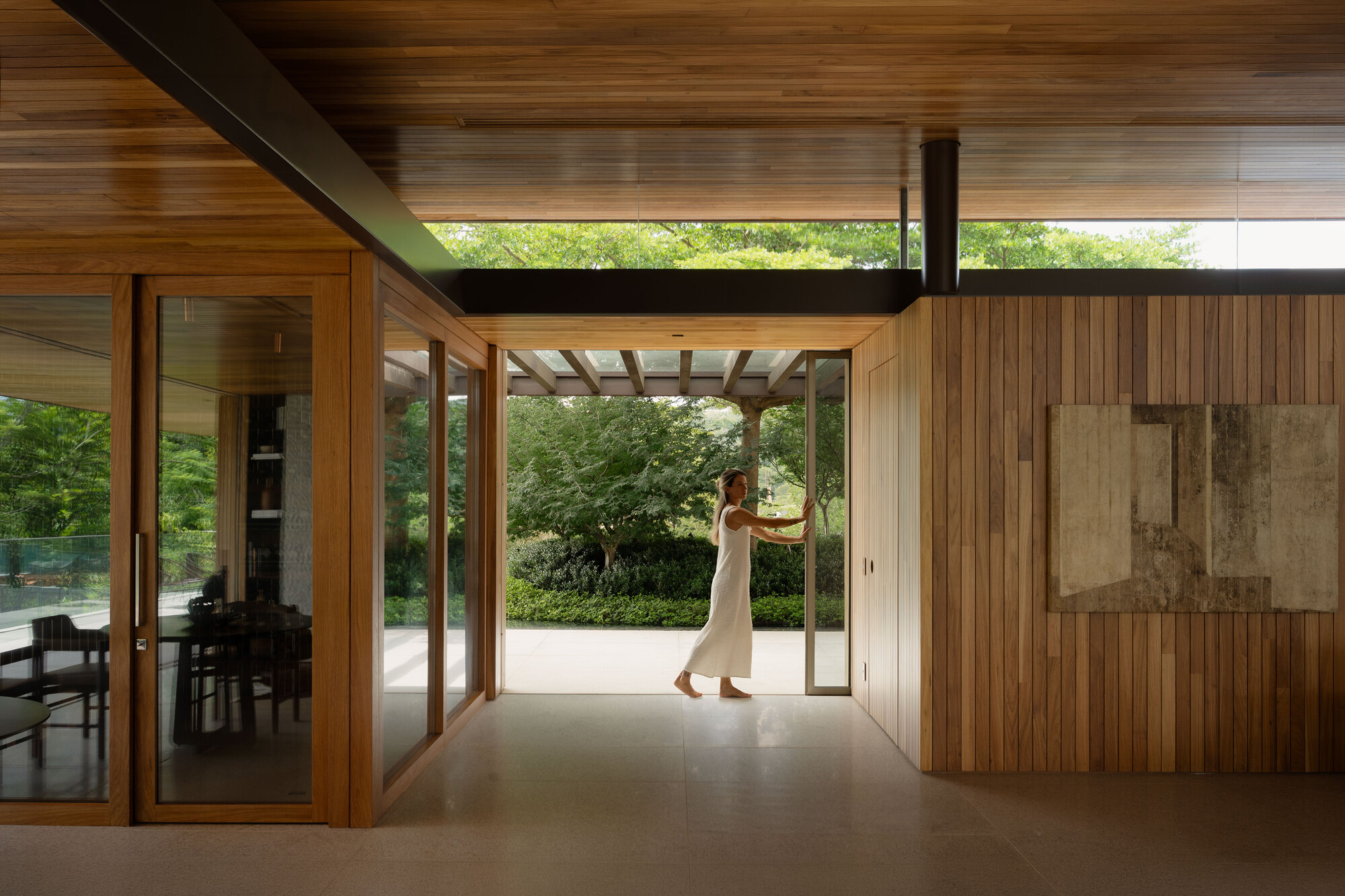 Woman in a white dress pushing open a glass door in a timber-lined interior that looks onto a green garden