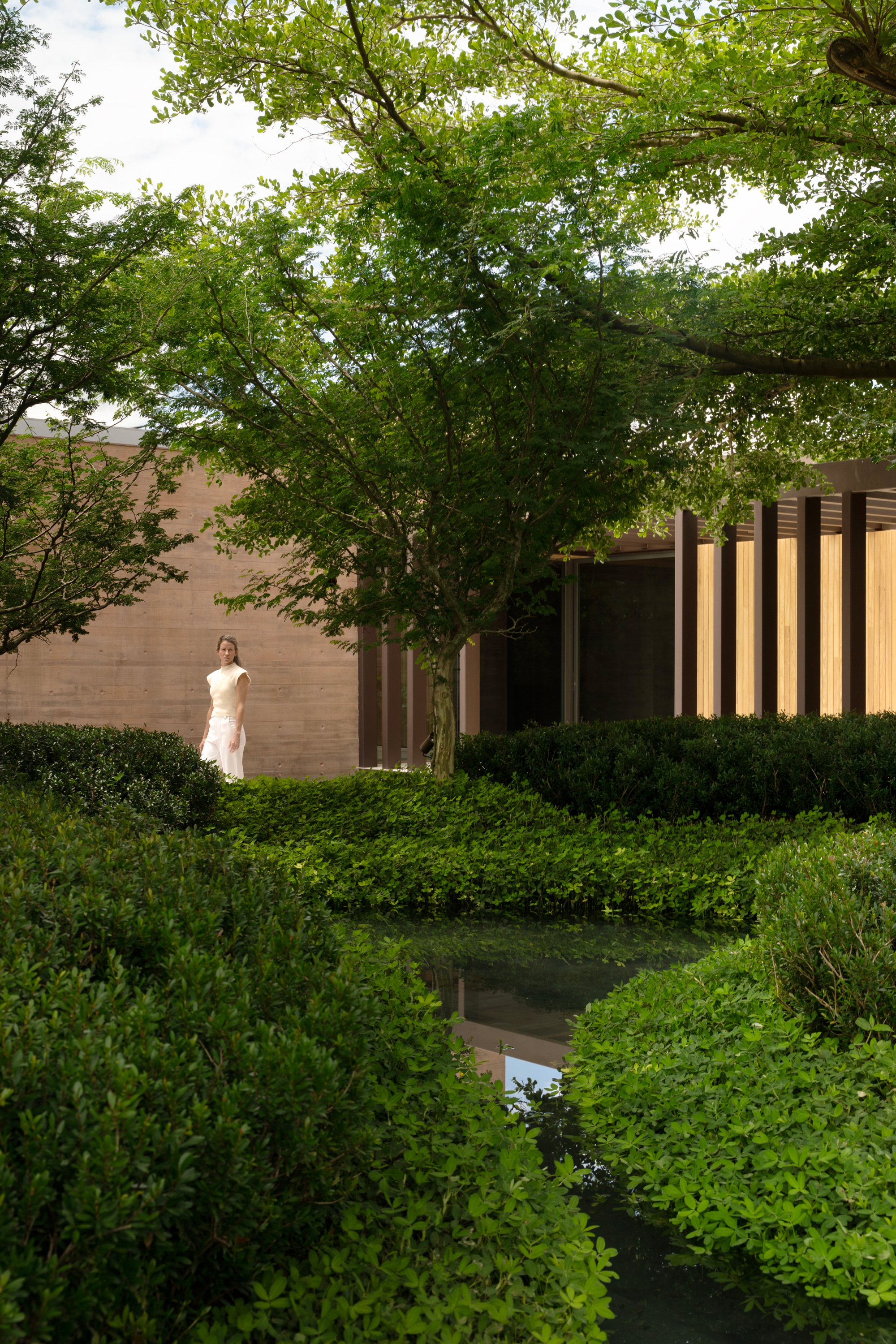 Woman standing beside a timber-screened pavilion and stone wall, seen across a reflecting pool and clipped shrubs under a leafy canopy