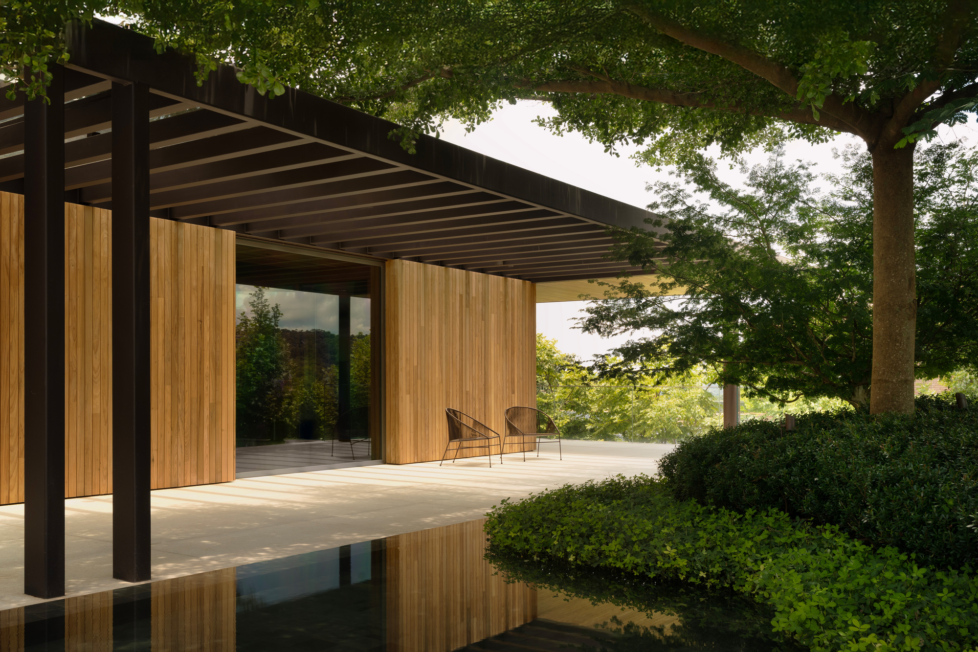 Covered terrace with dark steel columns, timber slat panels and two chairs, reflected in a pool beneath a spreading tree