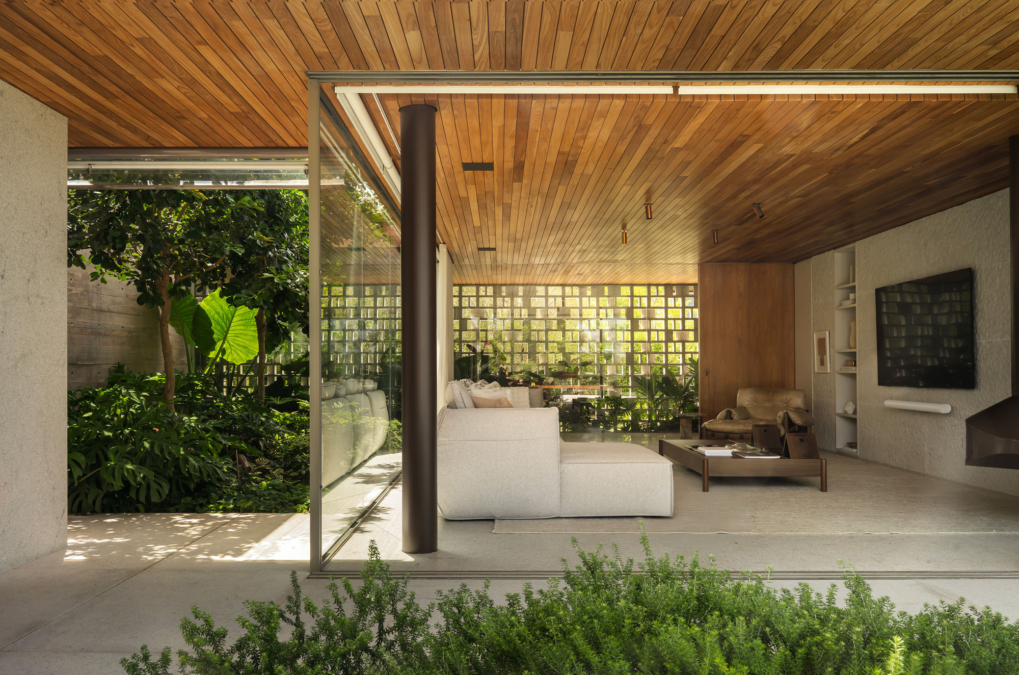 Living area opening onto a garden through frameless glass, with a timber ceiling, round column and perforated block wall beyond