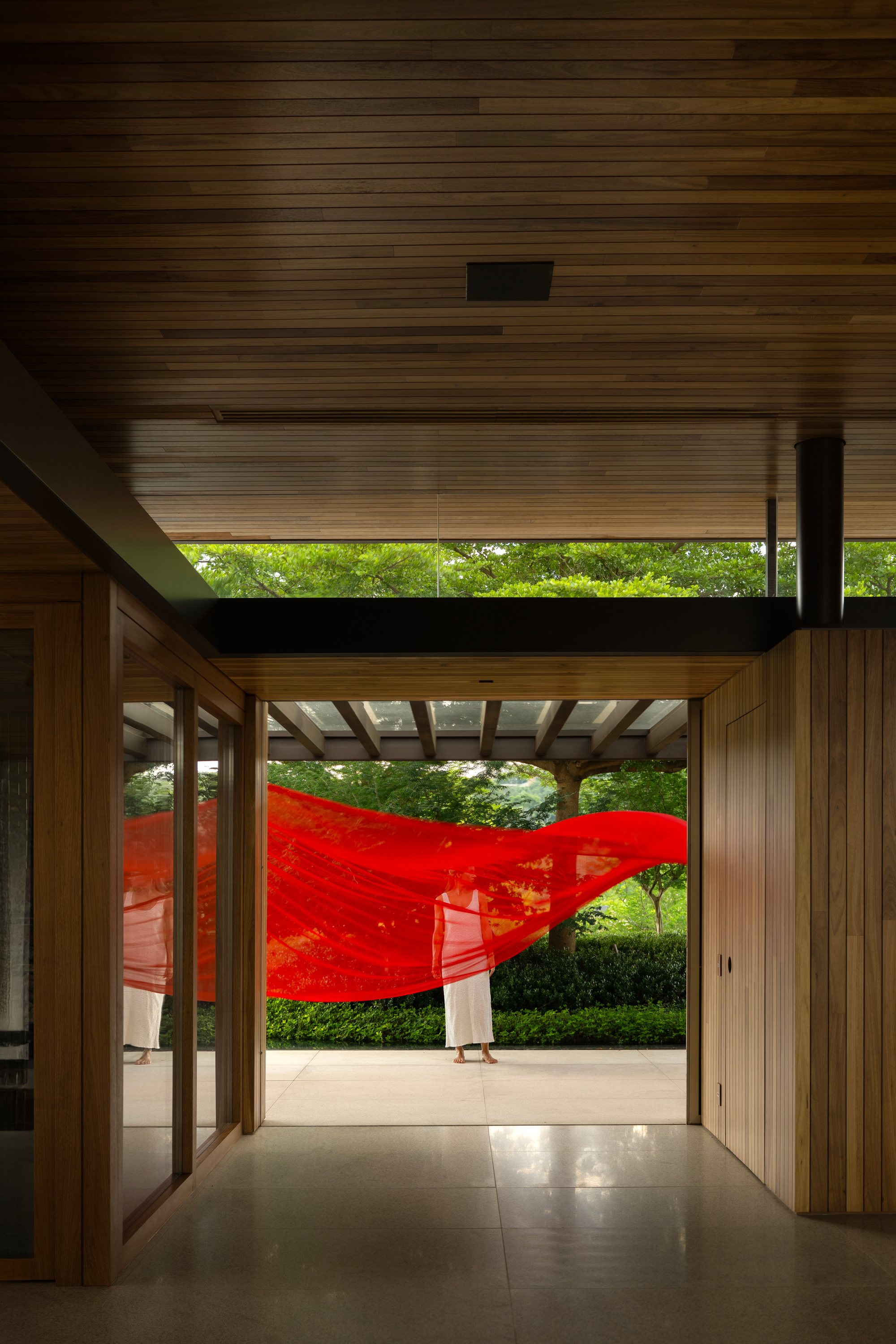 Red fabric billowing in the air as a figure stands on the terrace beyond a timber-lined entry and polished terrazzo floor