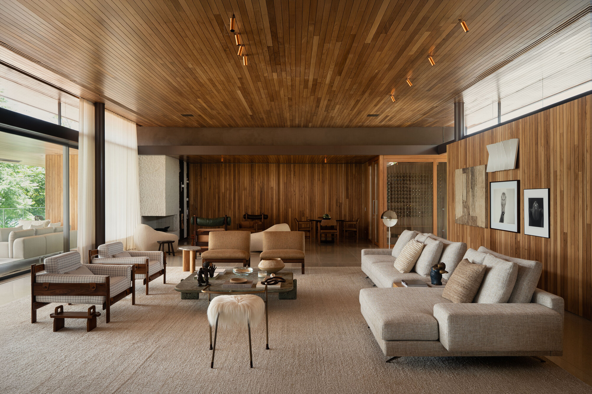 Long living room with timber-slat ceiling and wall cladding, pale sofas, leather armchairs and a wool rug in soft daylight