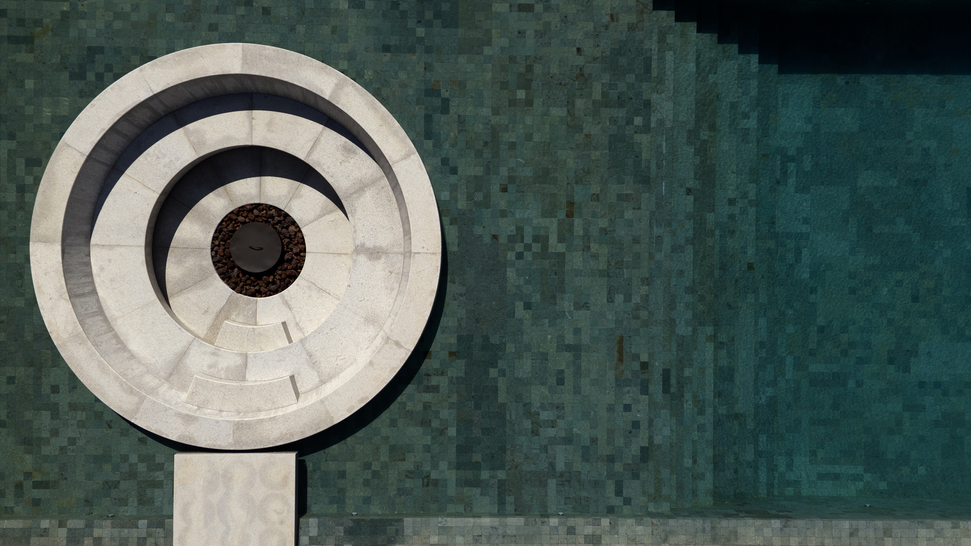 Top-down view of a circular stone fire pit with concentric steps set against green mosaic pool tile in hard sunlight