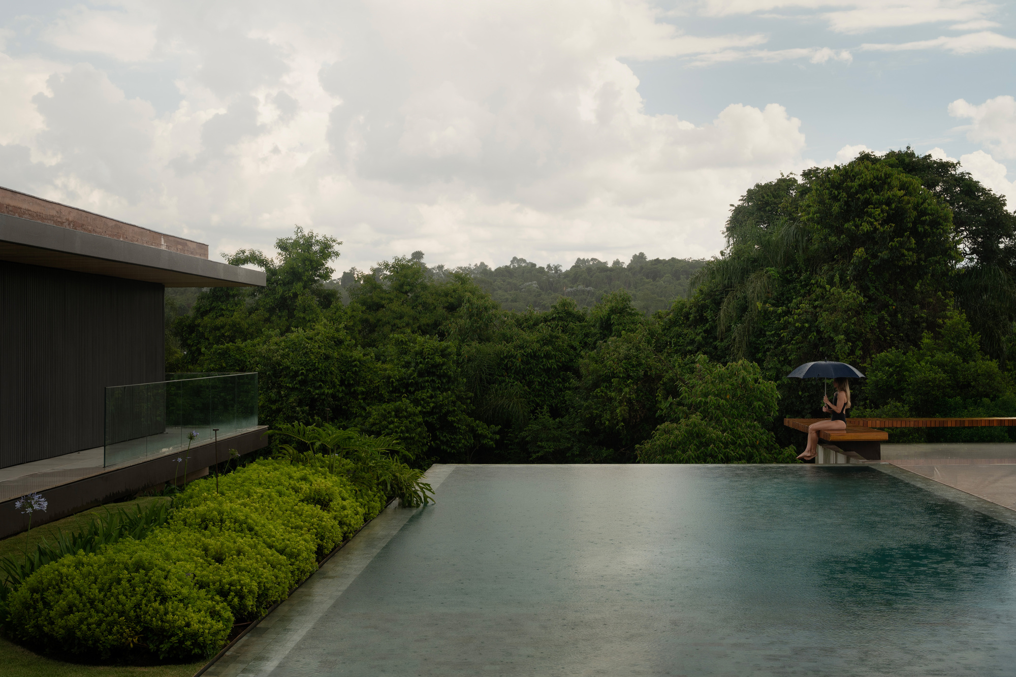 Infinity-edge pool facing a forest canopy, with a seated figure under an umbrella in light rain beside a glass balustrade