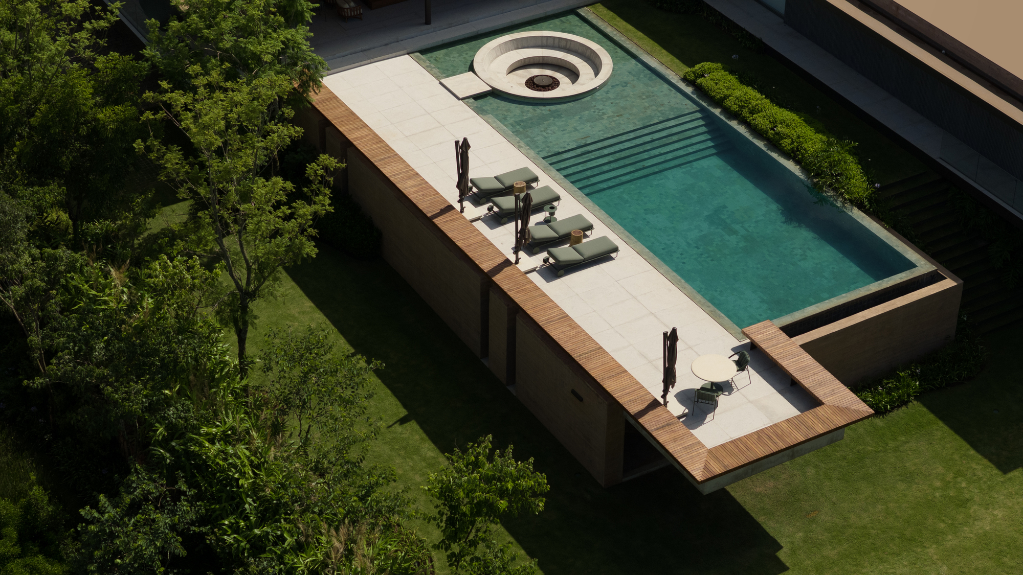 Aerial view of a rectangular pool with a round sunken fire pit, timber-edged deck and lounge chairs casting long shadows on lawn