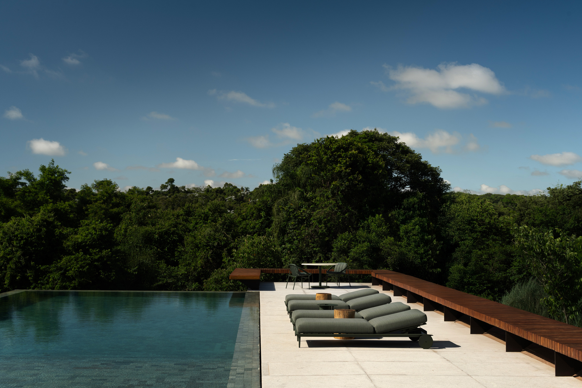 Pool deck with grey-green loungers and a long timber bench, facing dense forest under a blue sky with scattered clouds