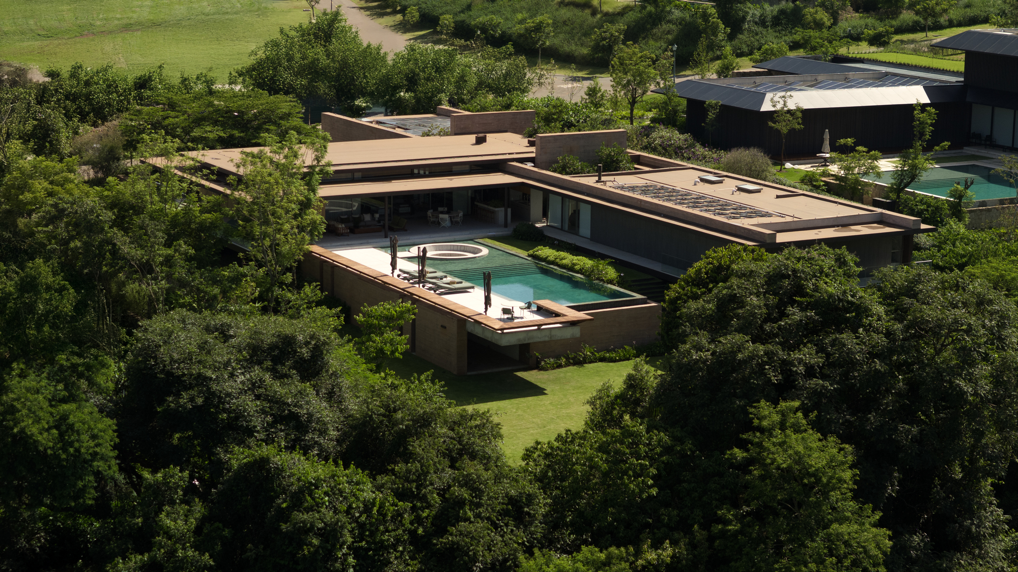 Aerial view of low flat-roofed pavilions with a pool and fire pit, surrounded by dense tree canopy in afternoon sun