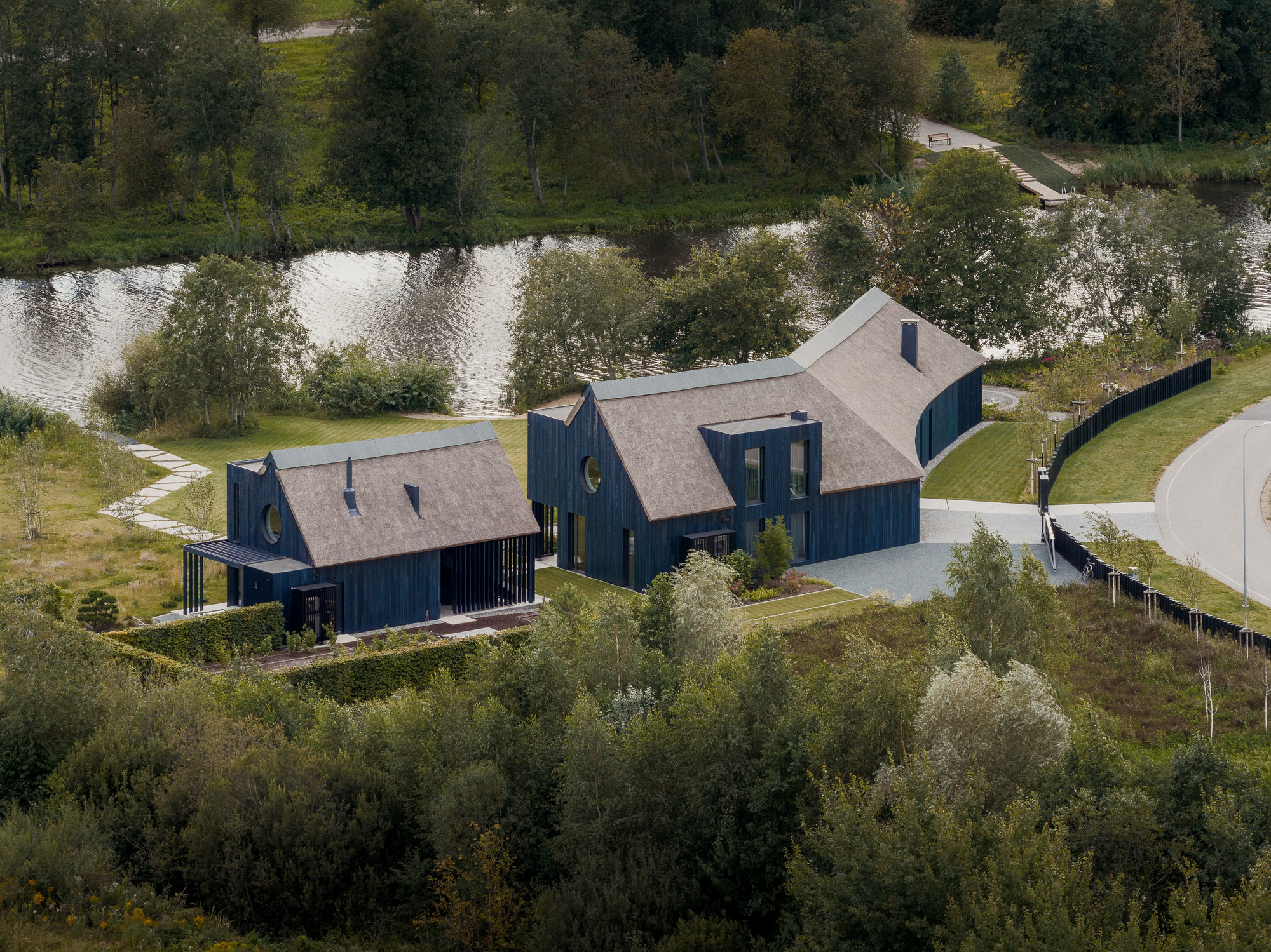 Aerial view of two dark timber houses with thatched roofs beside a river, ringed by trees on a grey summer day