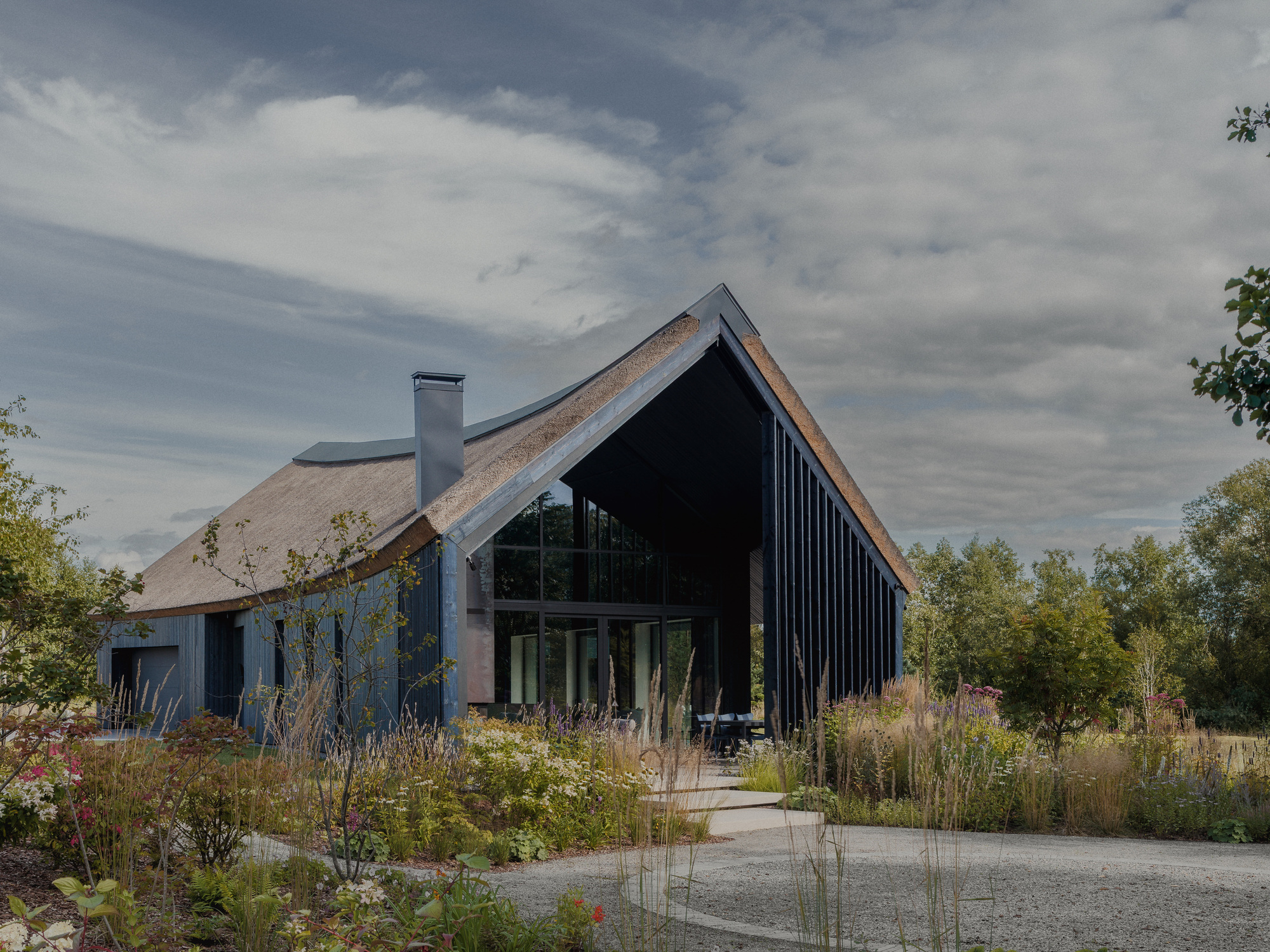 Gabled thatched house with a glazed end wall and chimney, set behind wildflower and grass planting under heavy clouds