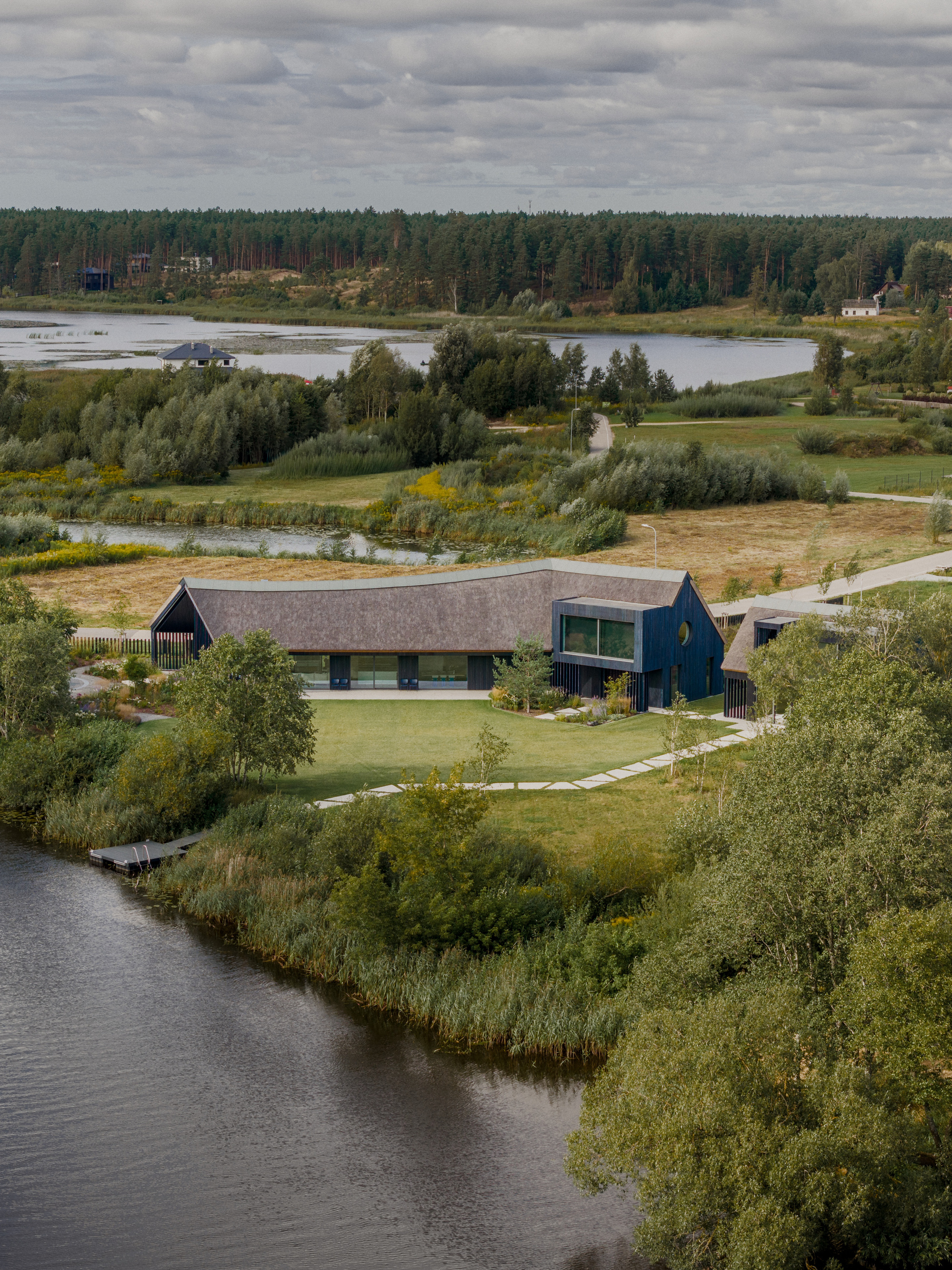 Drone view of the low thatched roof with a dark timber tower beside a lake, with a stepping-stone path across the lawn