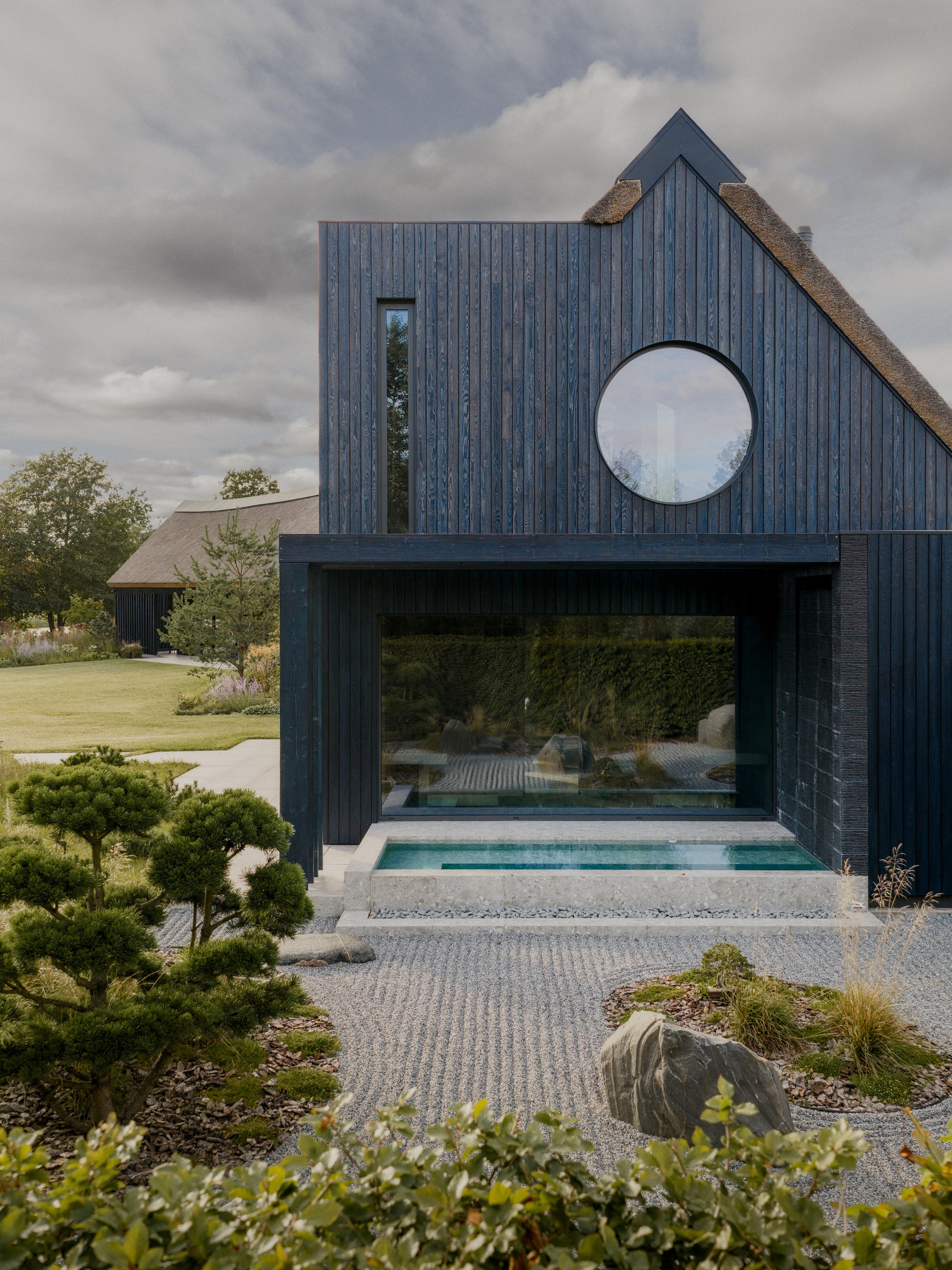 Charred timber gable with a round window above a glazed opening onto a small pool and raked gravel garden with a boulder