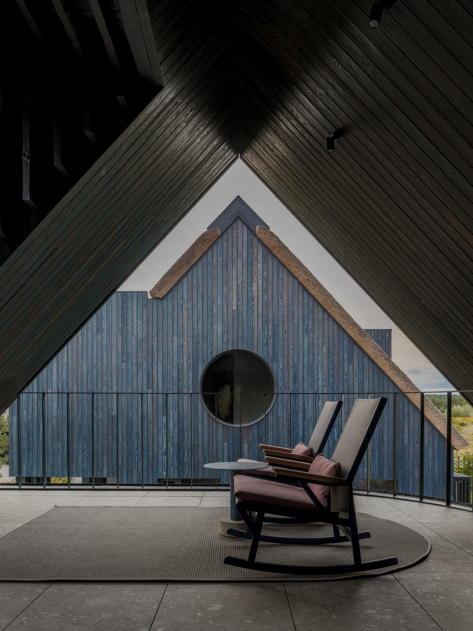 Rocking chair and round side table on a covered terrace framed by a dark timber ceiling, facing a gable with a circular window