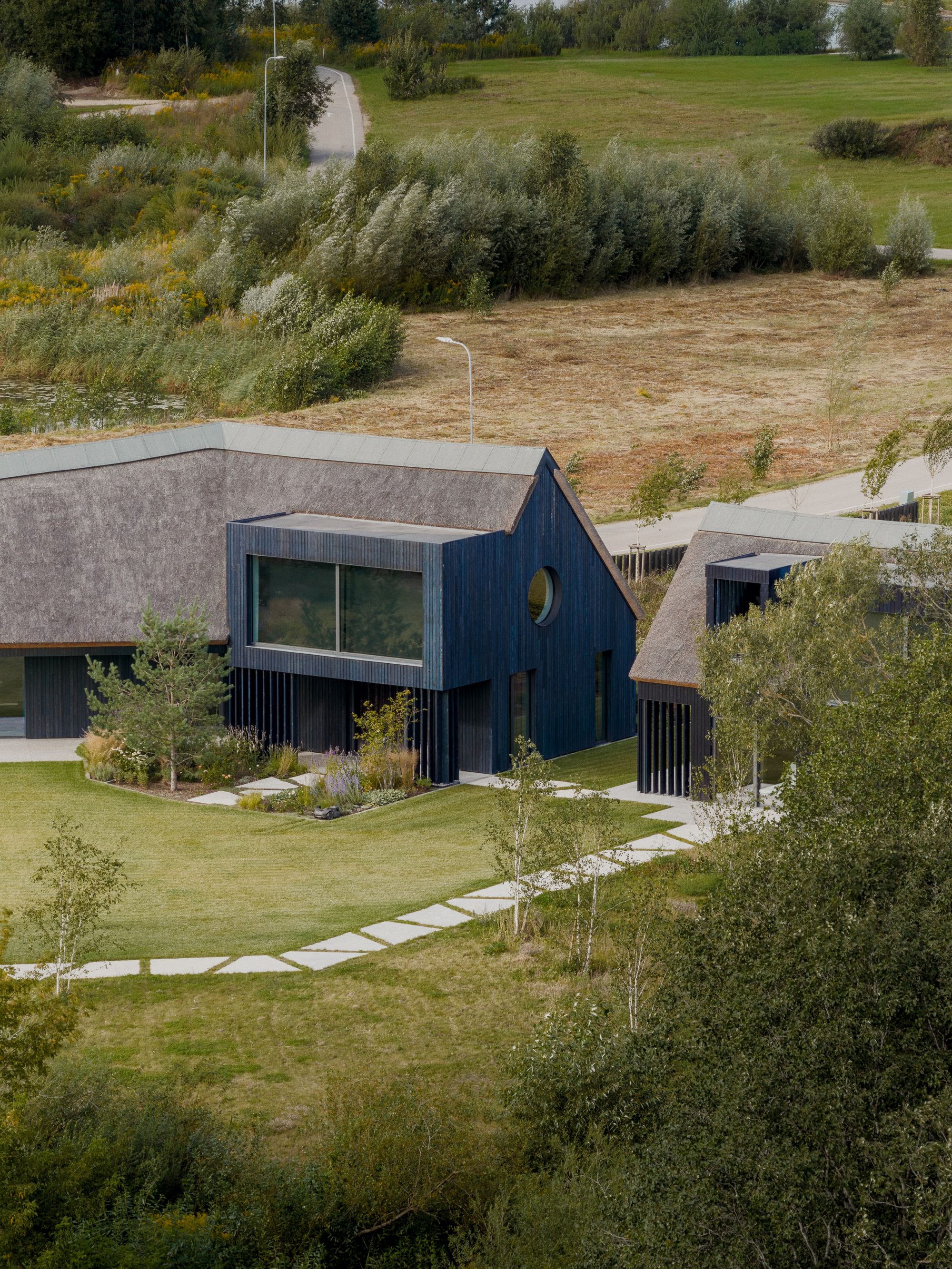 Aerial view of dark timber-clad house with thatched roof and stepping stone path across a lawn in late summer light