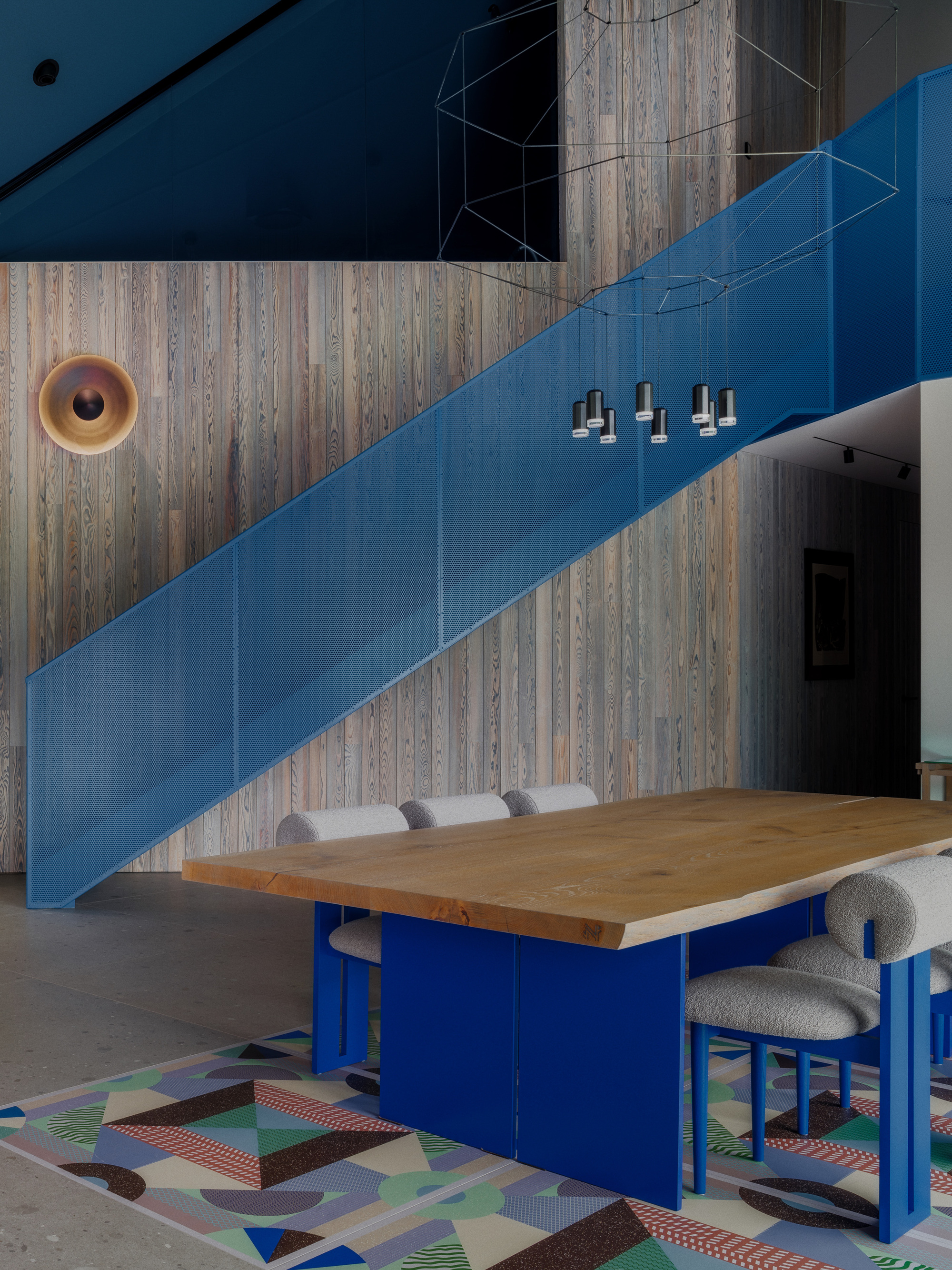 Blue perforated metal staircase above an oak dining table with blue legs, set on a geometric patterned rug