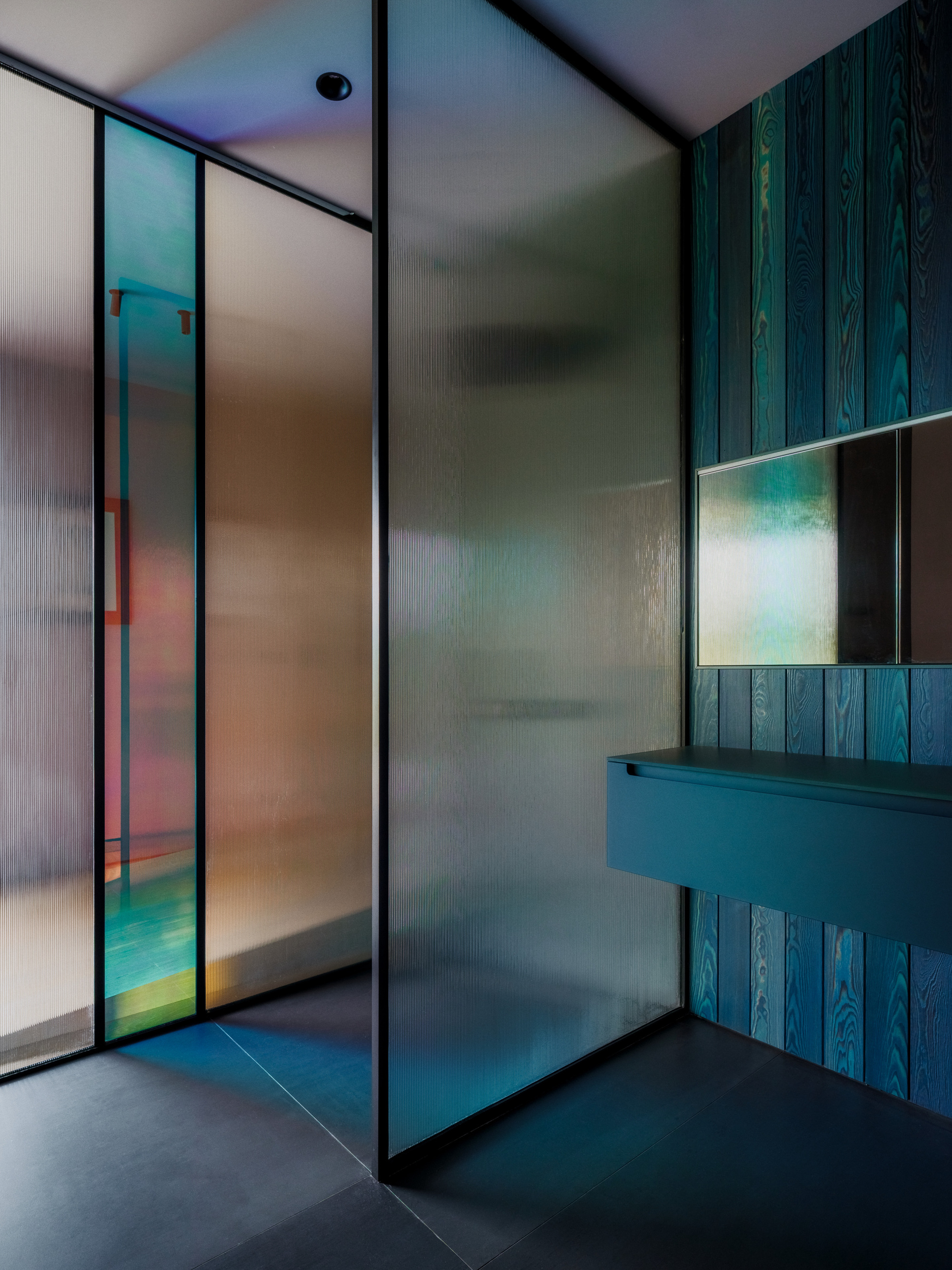 Fluted glass partitions with iridescent panel beside a teal-stained timber wall, floating vanity and mirror in a dim bathroom