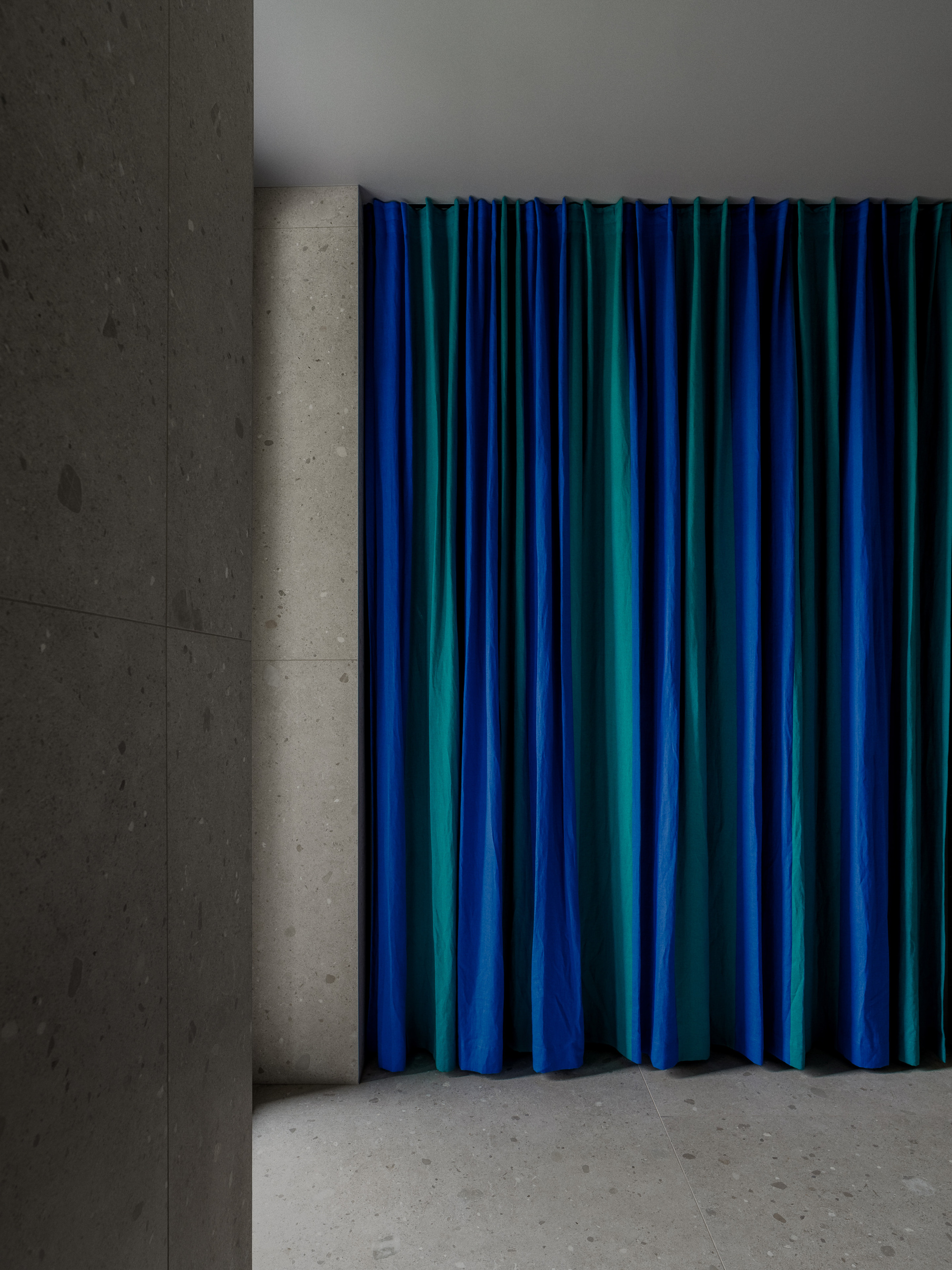 Floor-to-ceiling blue and green striped curtain beside speckled terrazzo-style stone wall panels and floor in soft grey light