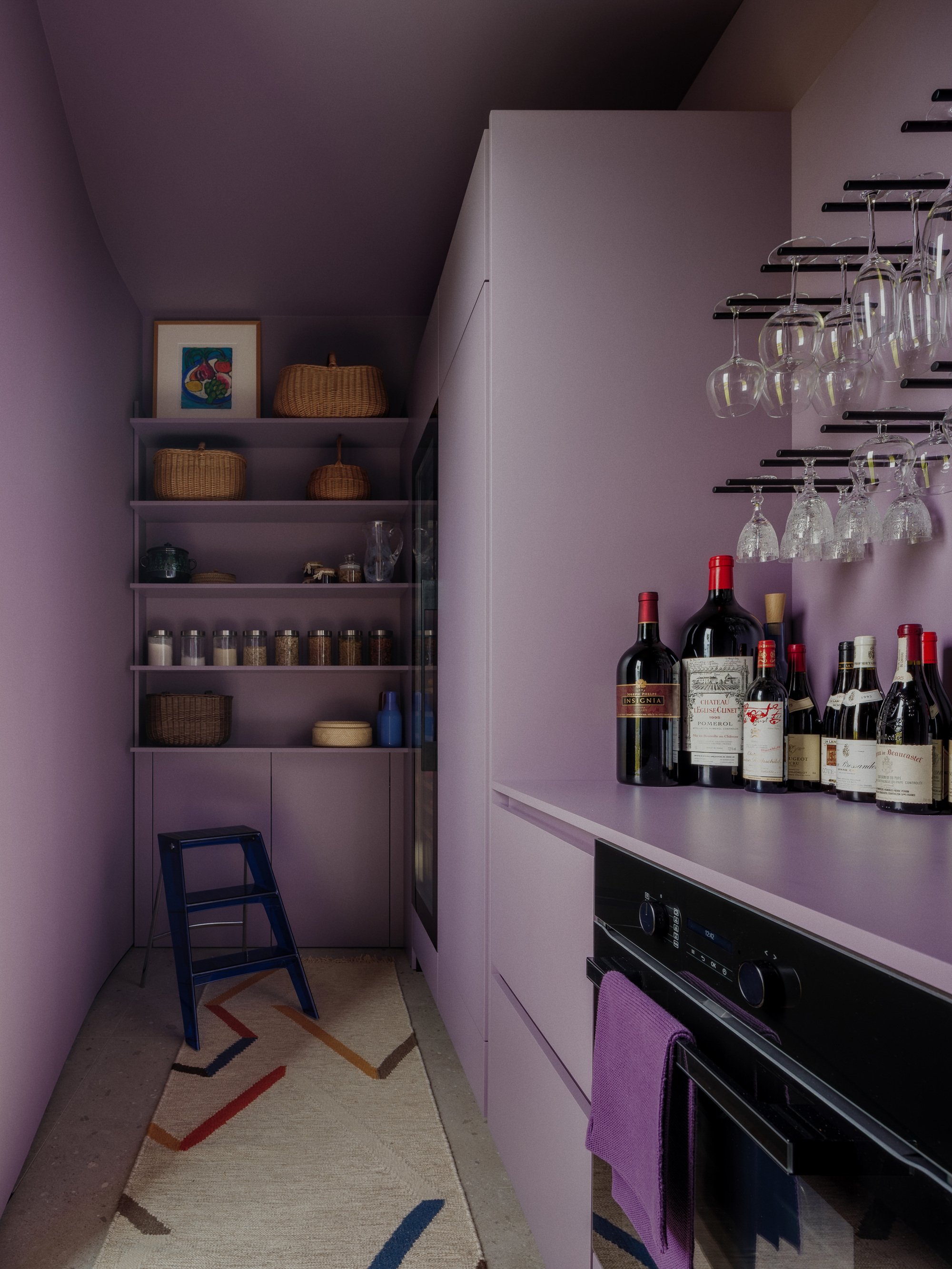 Narrow lilac pantry with open shelving, wicker baskets, a blue step stool, hanging wine glasses and bottles on the counter