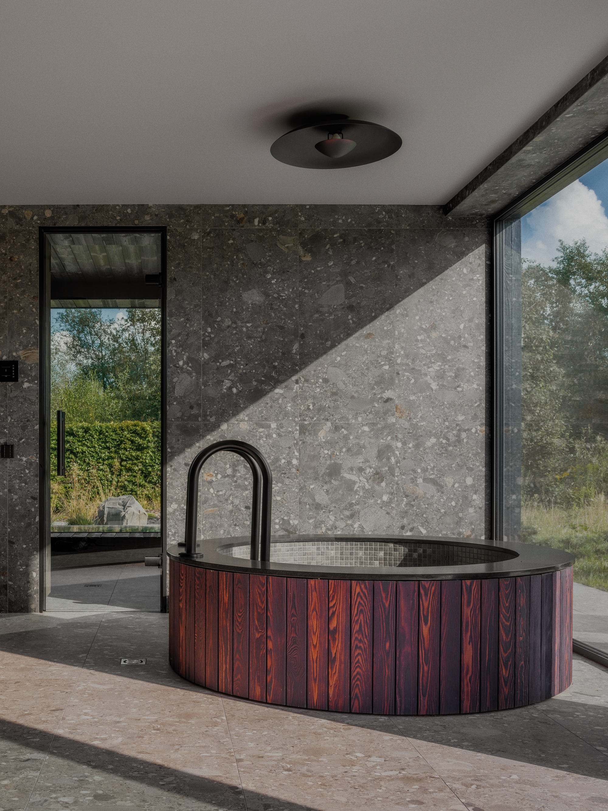 Round tub clad in charred red timber slats against a terrazzo-like stone wall, with sunlight cutting across the floor