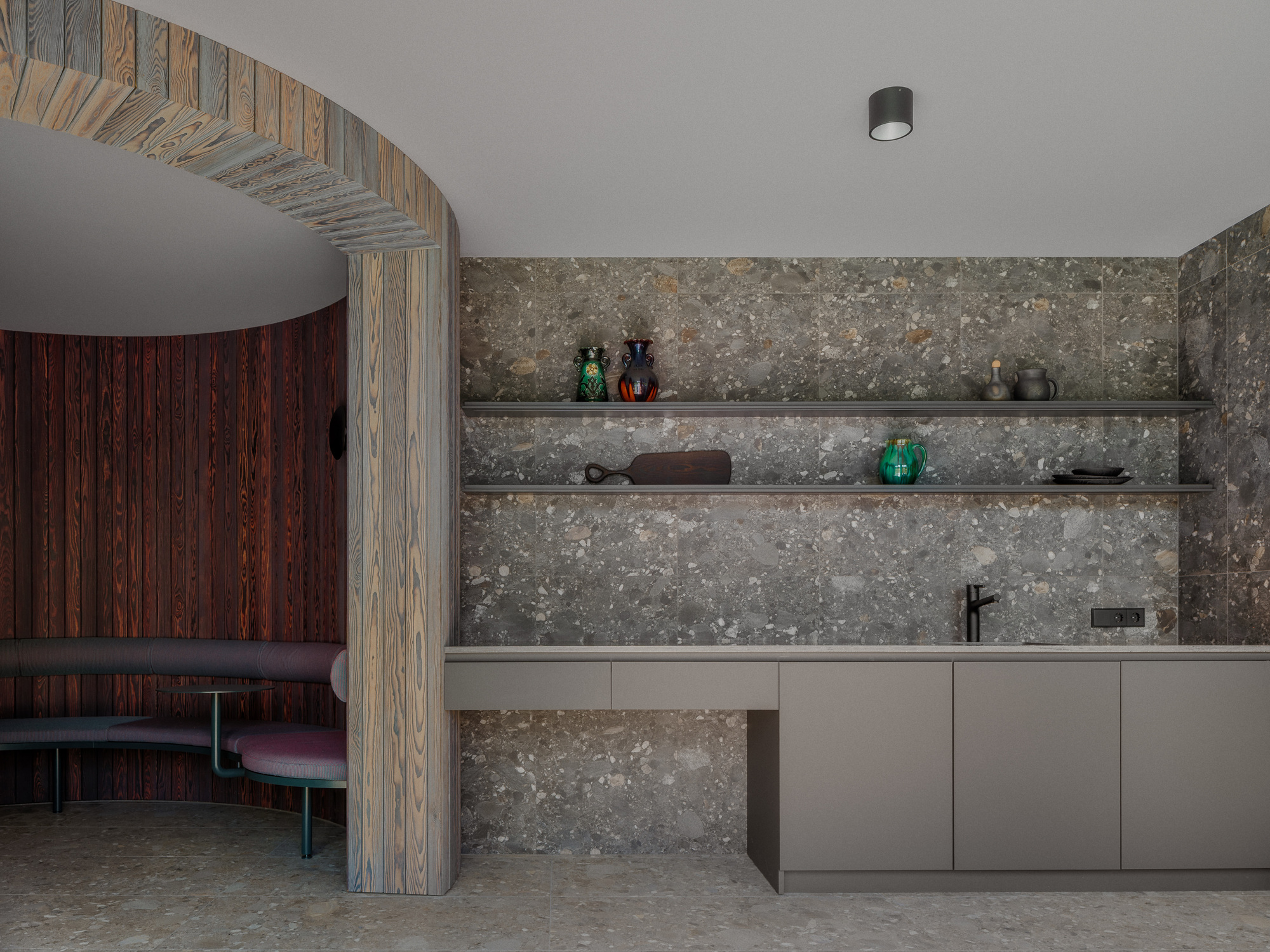 Stone-clad kitchenette with floating shelves holding ceramics, beside a timber arch opening onto a curved upholstered bench