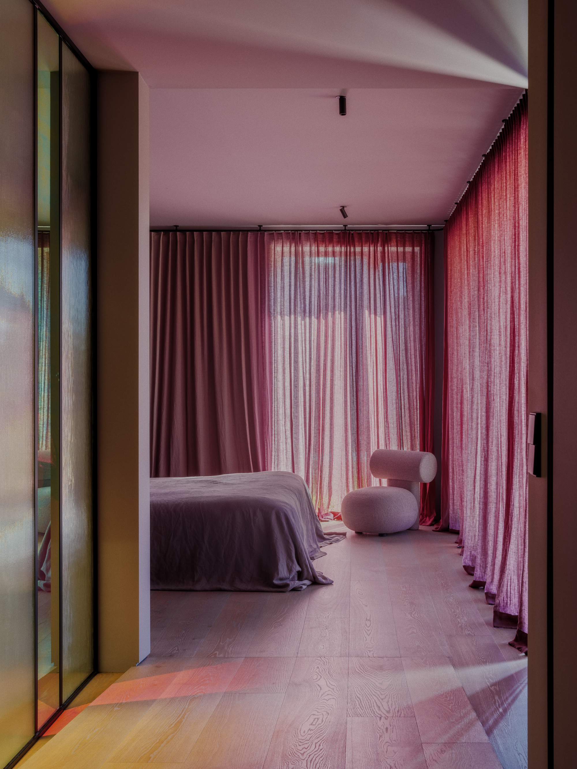 Bedroom with sheer pink curtains, a low bed and a round white stool, lit by low colored sunlight