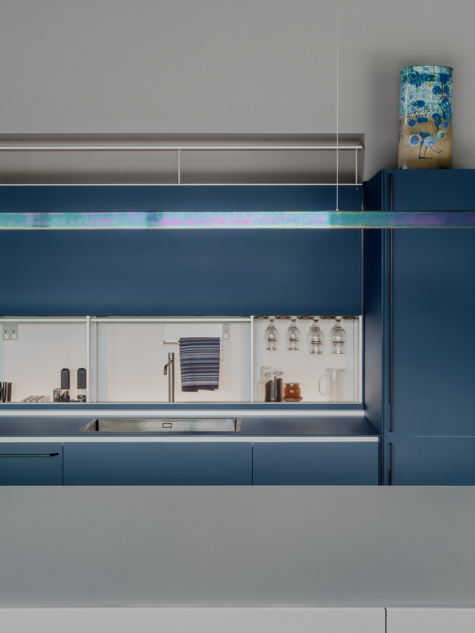 Blue kitchen run with steel sink, mirrored splashback and hanging glasses beneath an iridescent light bar