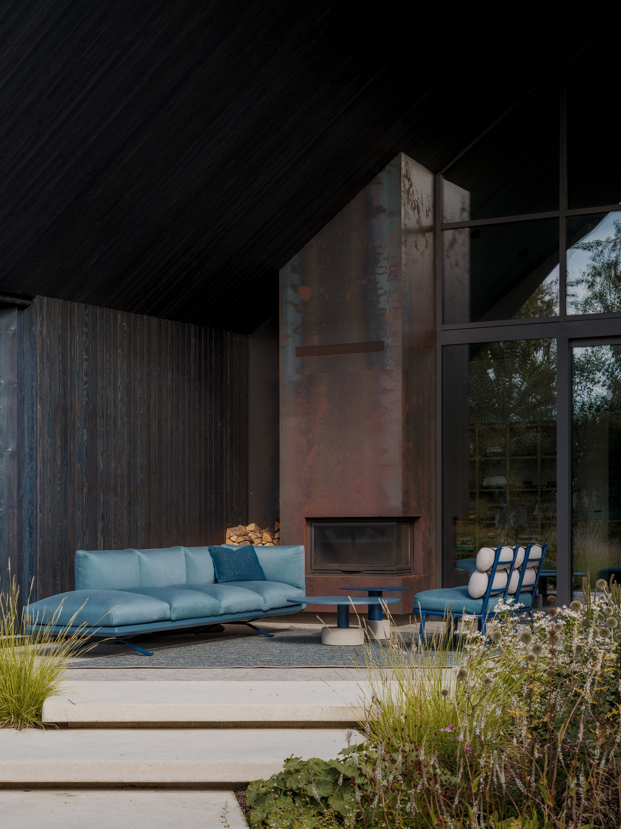 Covered terrace with a weathered steel fireplace, blue sofa and charred timber walls, edged by grasses and wildflowers