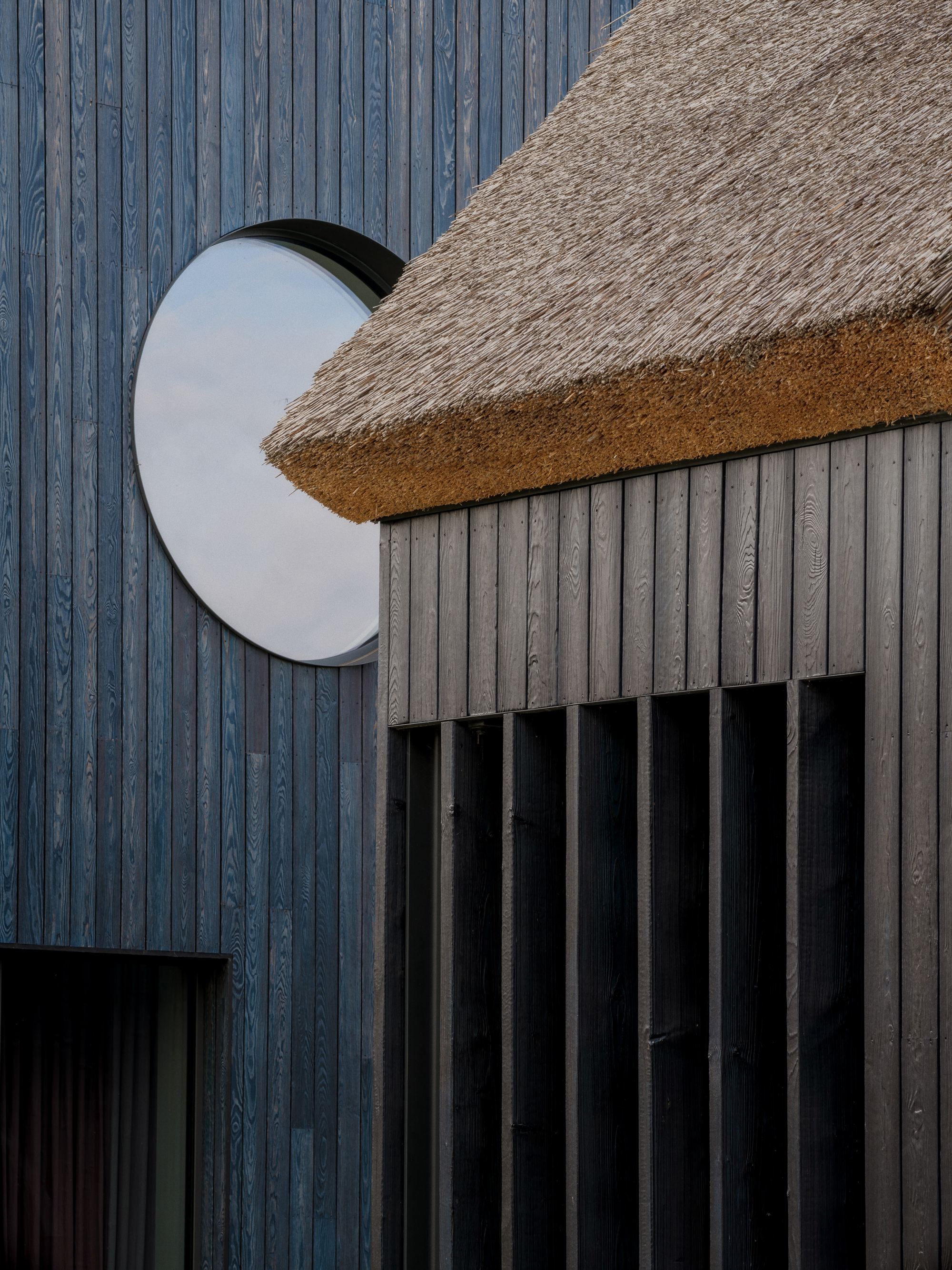 Close-up of a round window in blue-stained vertical boards beside a thatched eave over slatted timber screening