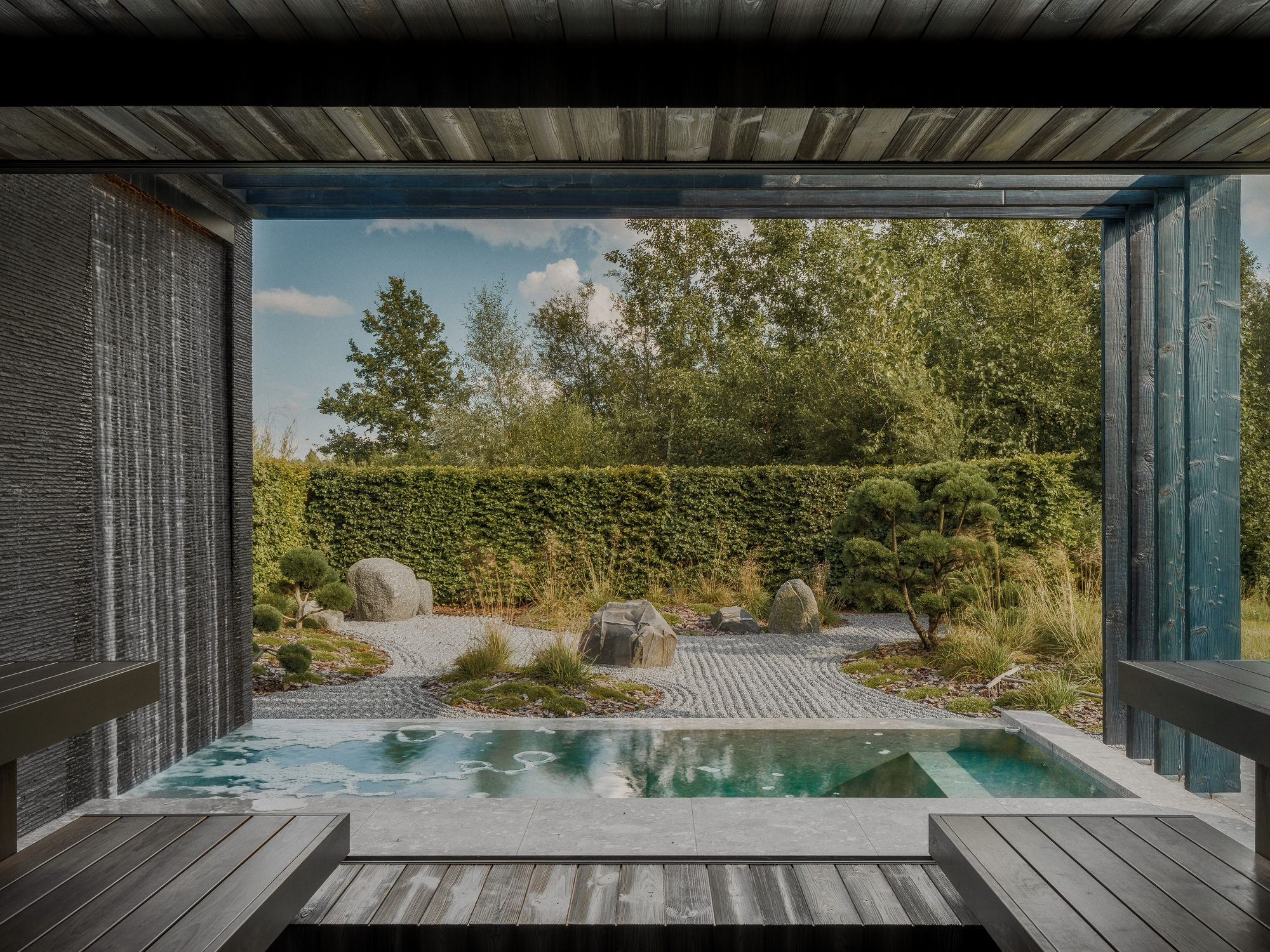 View from a timber-lined sauna pavilion across a small plunge pool to a gravel garden with boulders and clipped pines in summer light