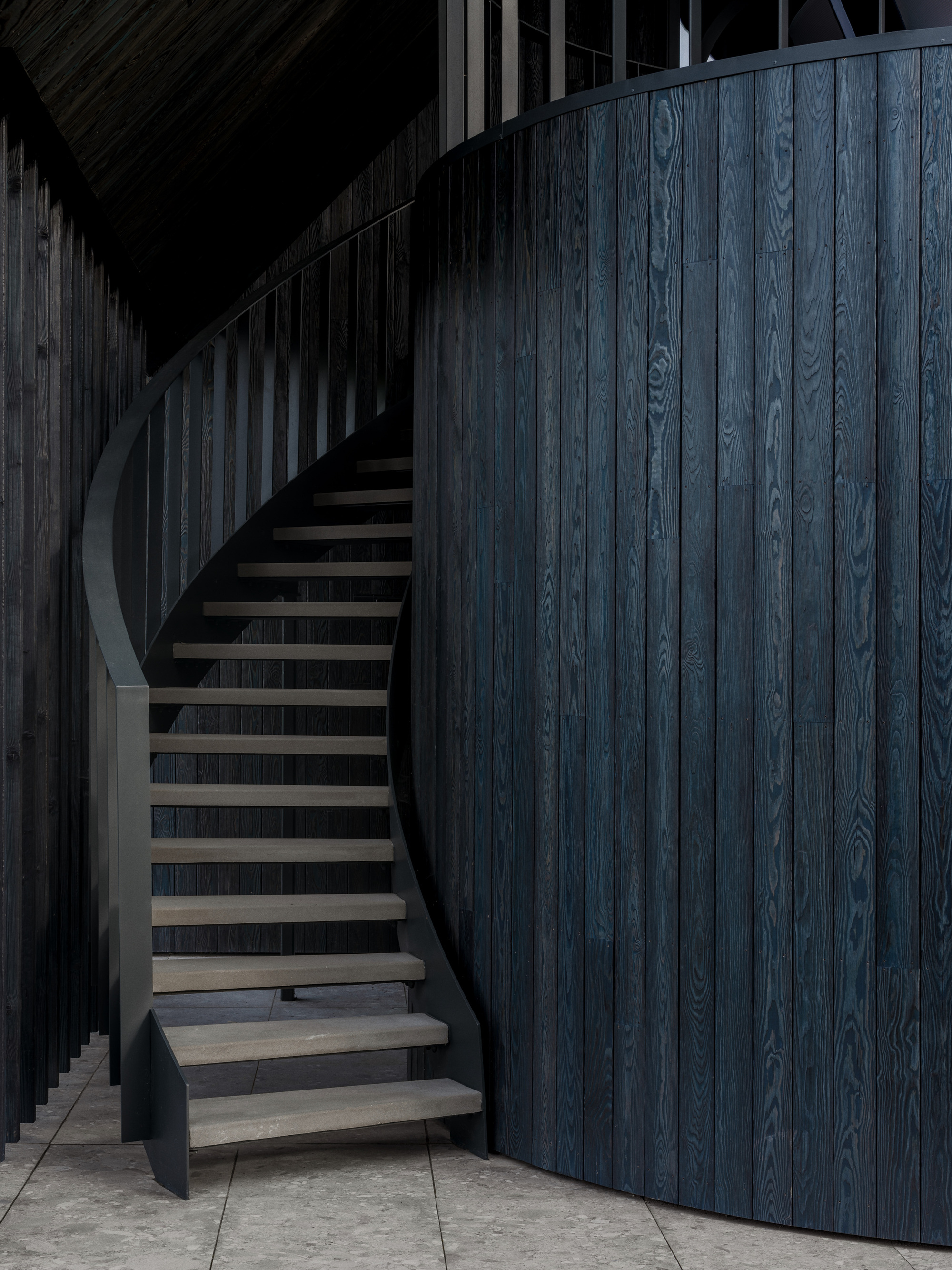 Curved stair with floating timber treads beside a charred dark blue wood wall, lit by soft low light