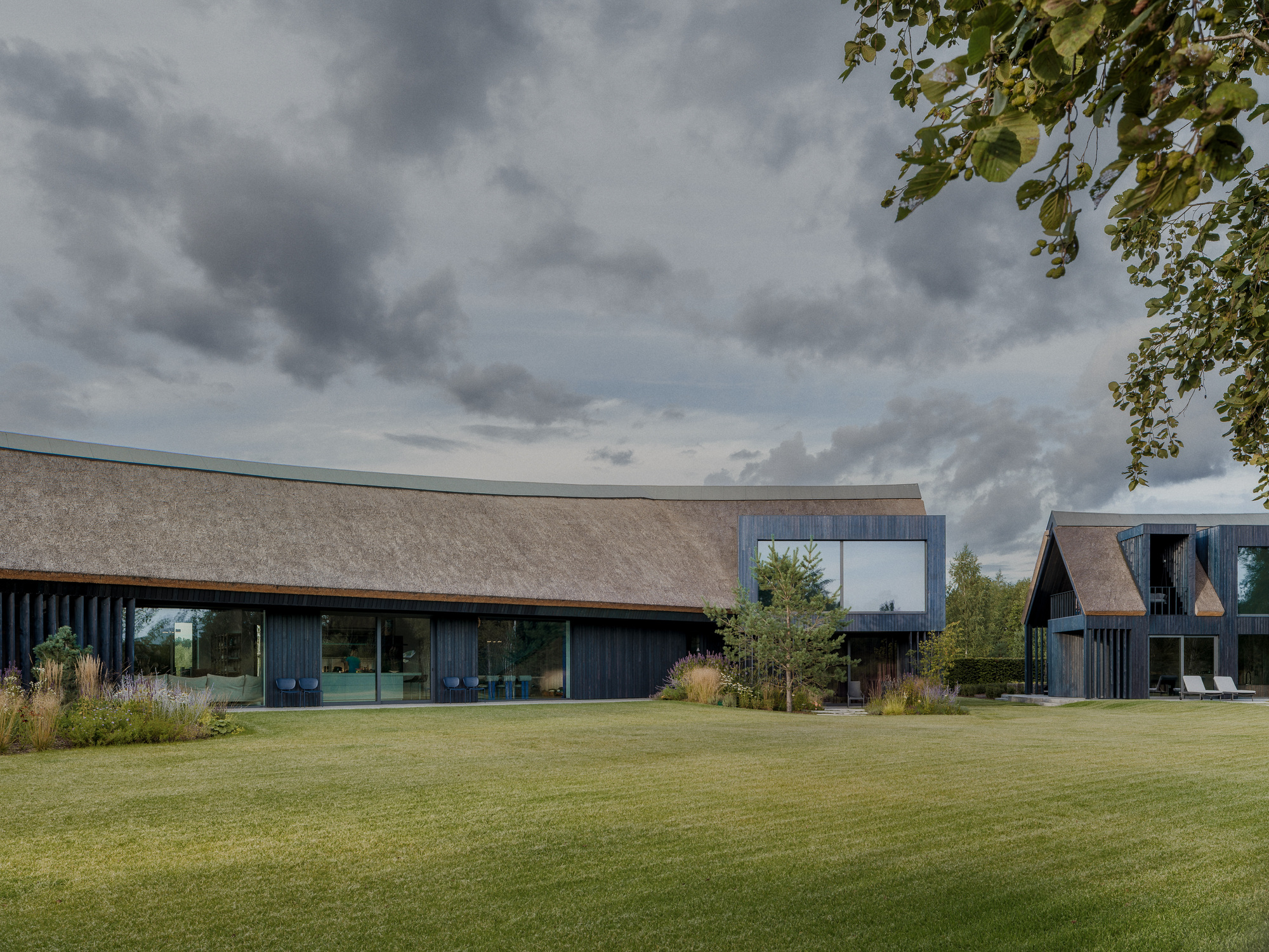 Long thatched roof over a glazed dark timber wing, with a gabled volume at right, seen across a lawn under heavy clouds