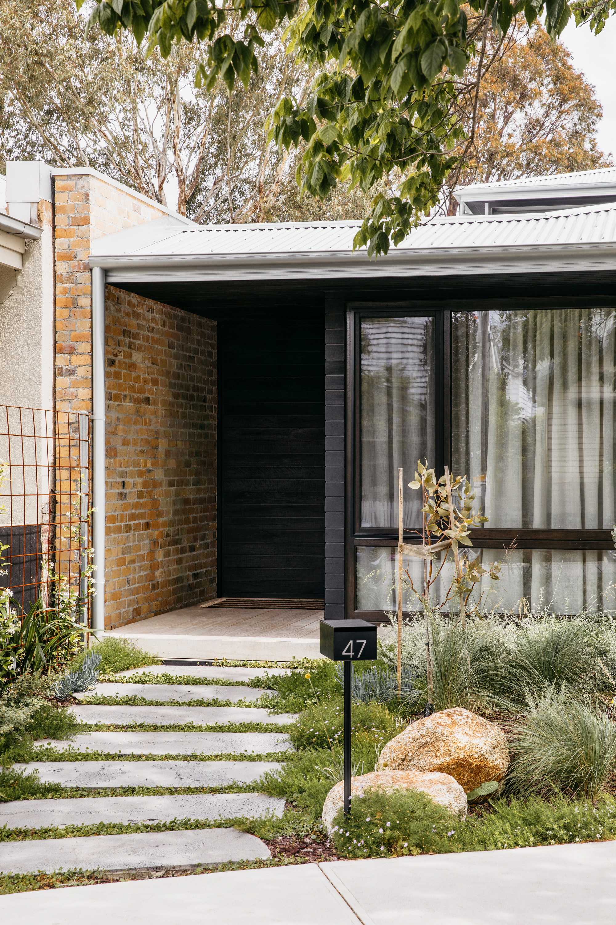 Entry porch with a dark timber door and recessed brick wall, stepping stones crossing a grassy planted garden with boulders
