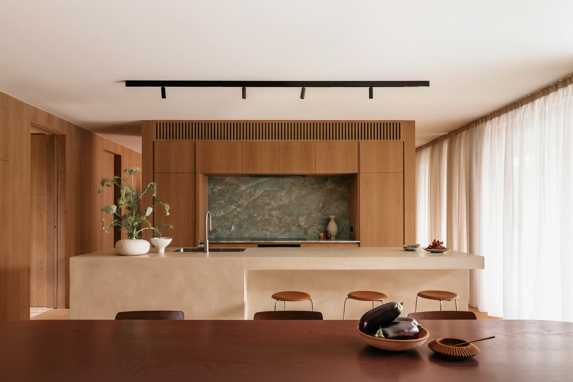 Kitchen island in pale plaster with a green stone splashback and timber joinery, sheer curtains drawn along the window wall