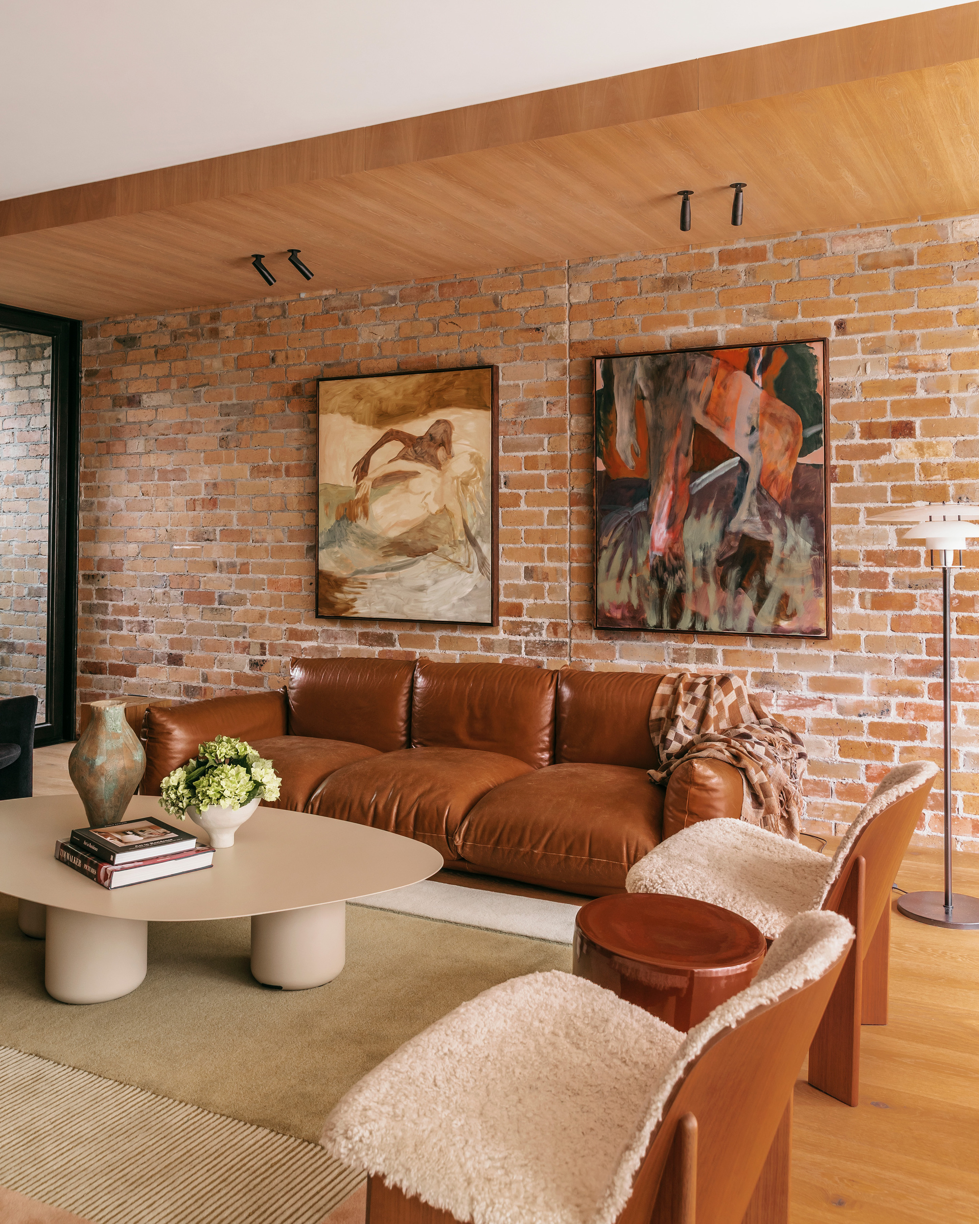 Leather sofa beneath two framed paintings on a brick wall, with sheepskin-covered timber chairs and an oval coffee table in daylight