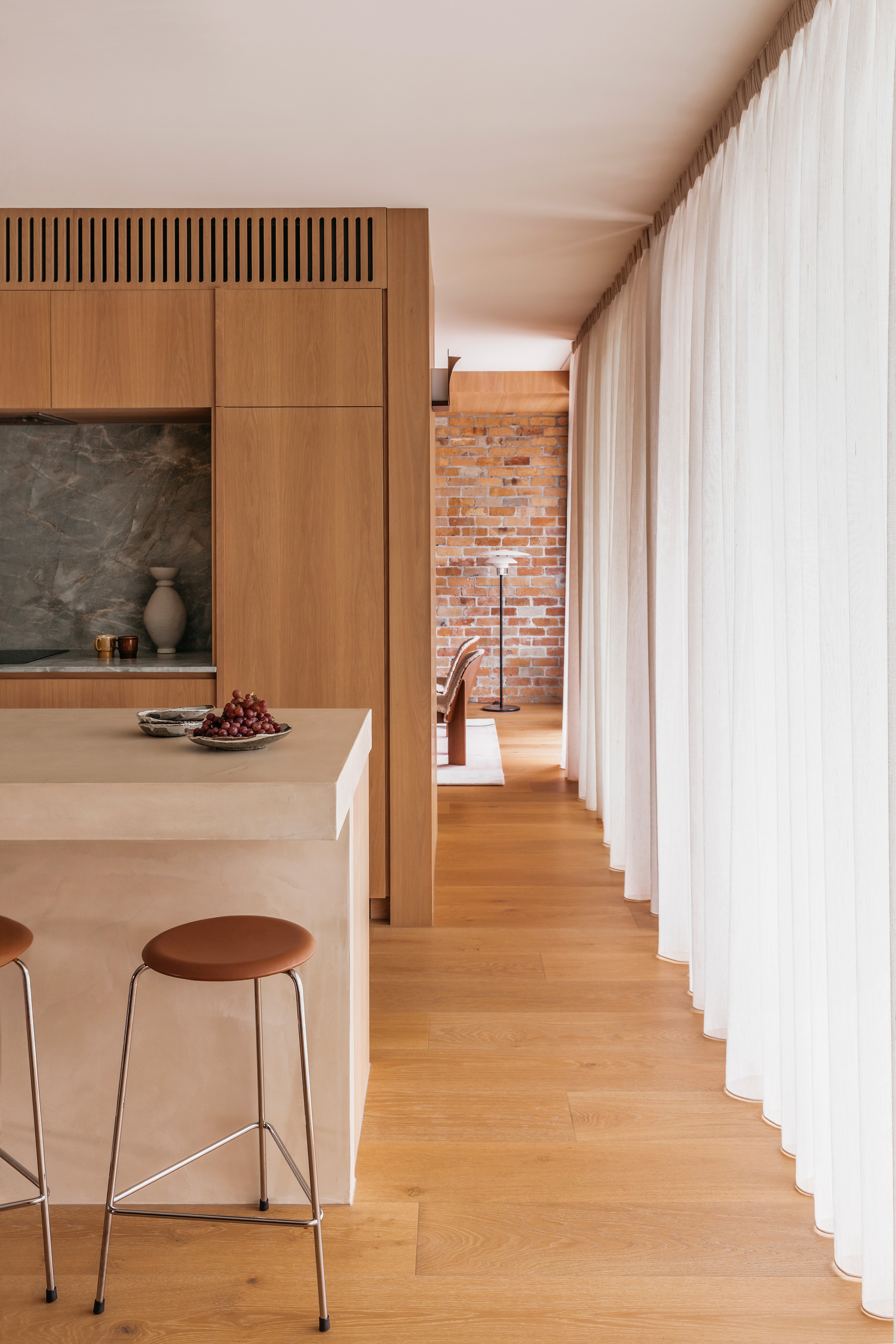 Side of the kitchen island with a leather stool, oak floor running past floor-length white curtains toward a distant brick wall