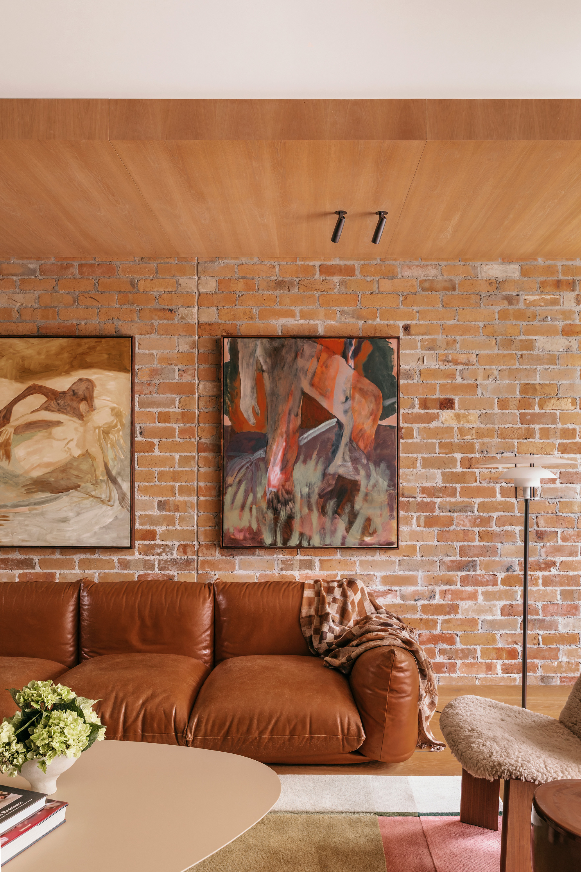 Tan leather sofa below two framed paintings on a reclaimed brick wall, with track spotlights on the timber ceiling
