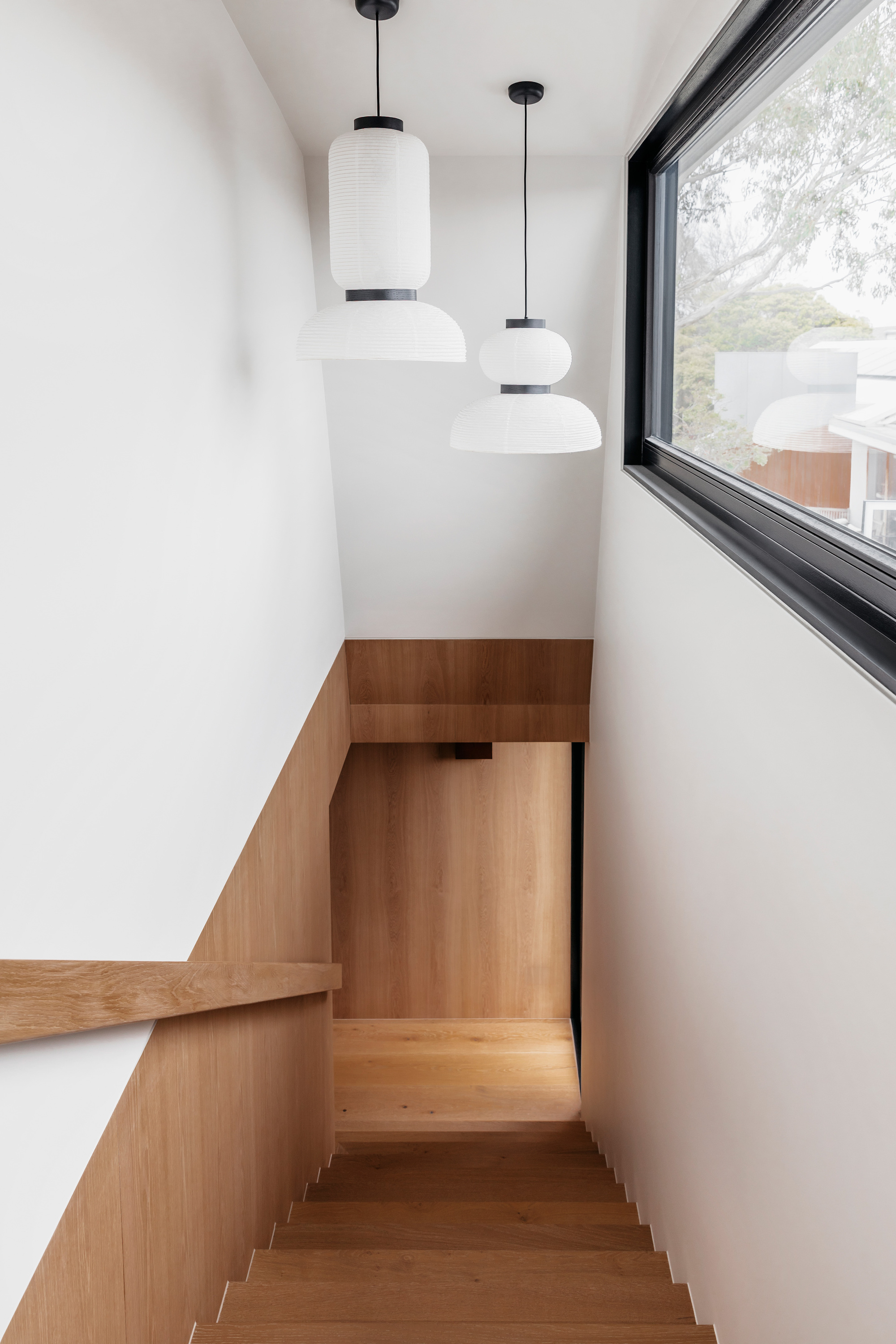 Staircase descending between white walls and oak panelling, with two paper pendant lights and a tree seen through the window