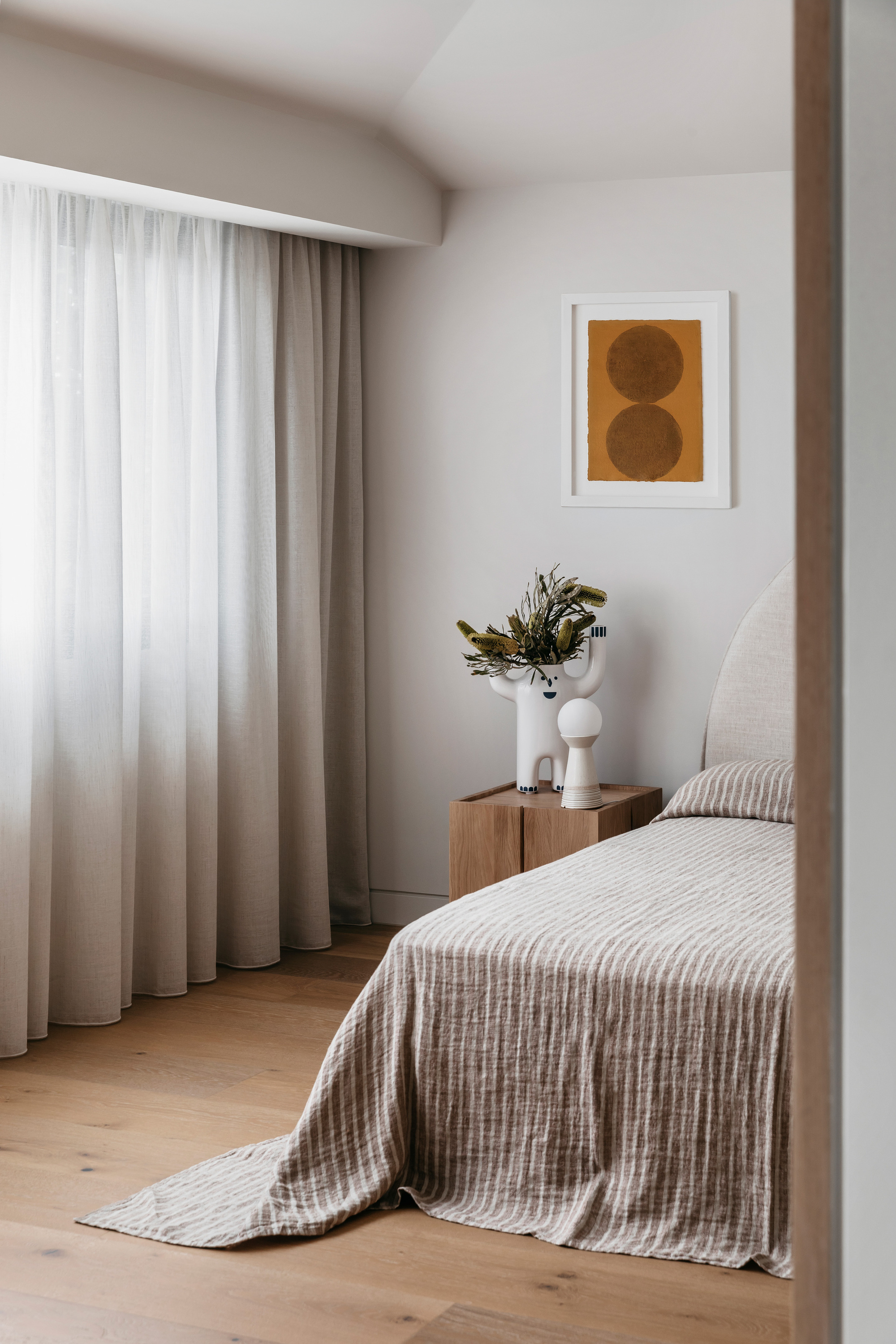 Bedroom corner with a linen-striped bedspread, timber side table and white vase of dried banksia, soft daylight filtering through sheer curtains