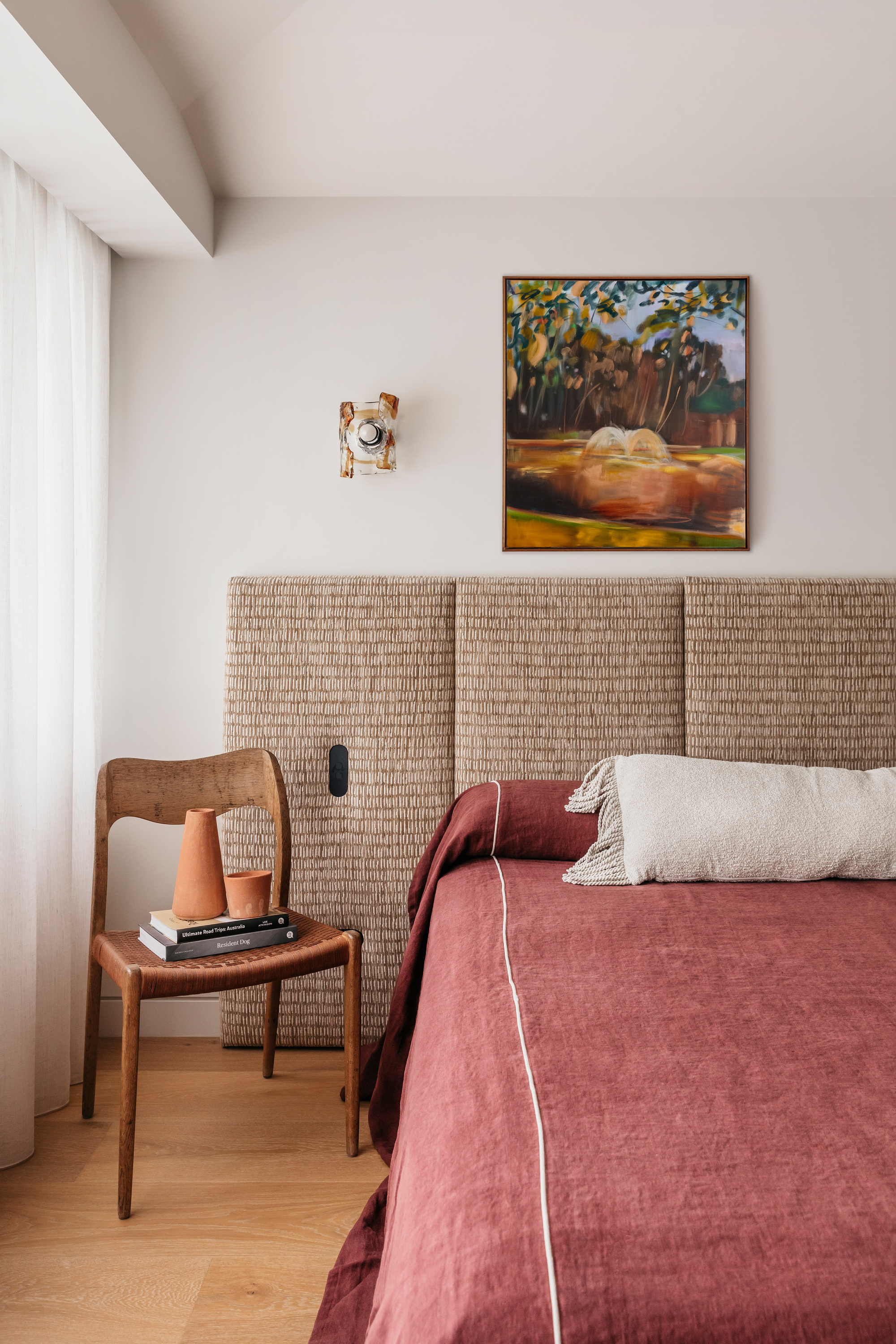 Woven panel headboard above a rust linen bed, with a timber chair holding terracotta vessels beside a sheer curtain