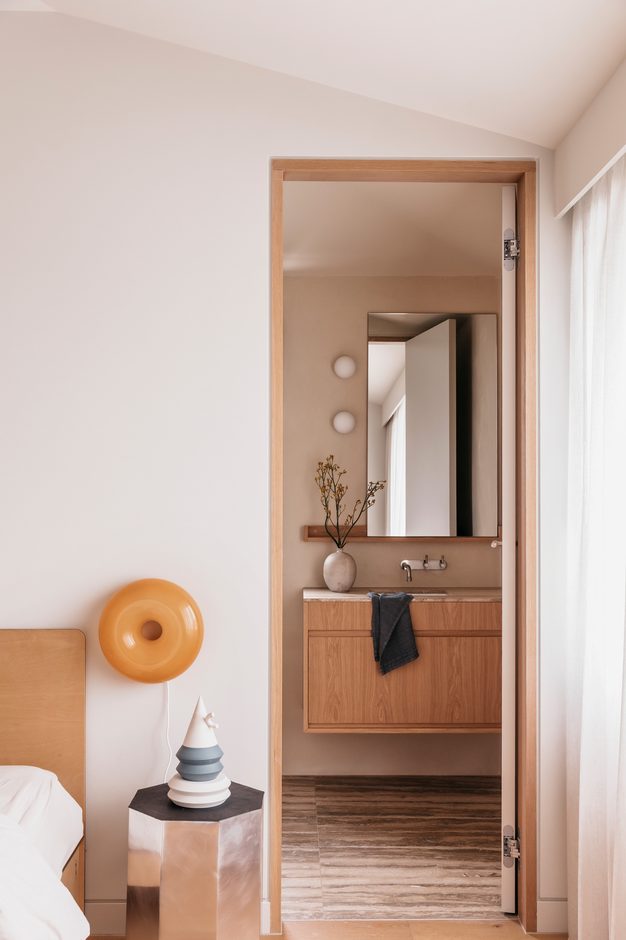 Doorway from the bedroom to an ensuite with floating oak vanity, travertine floor and a mirror, beside a round orange wall lamp